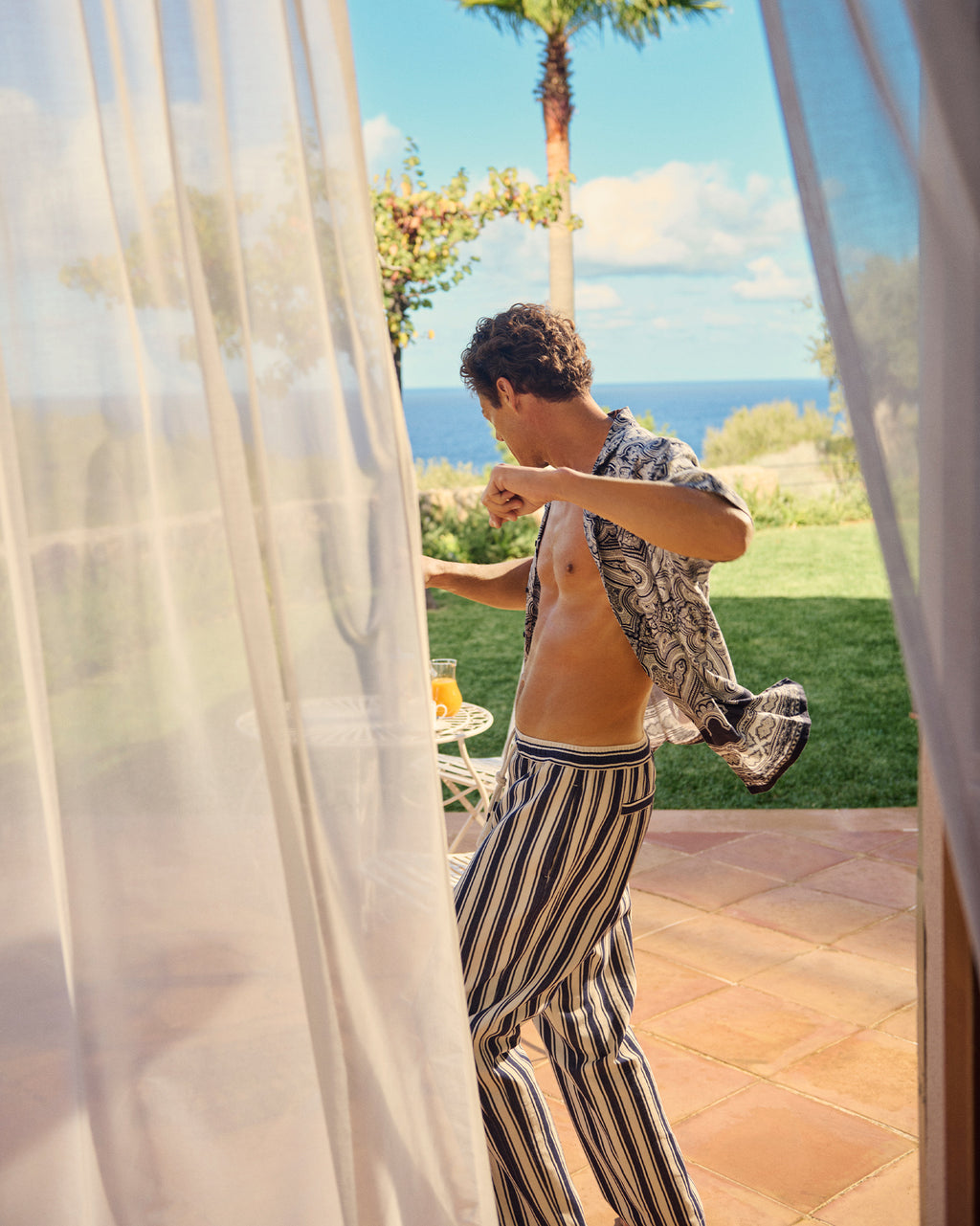Man in striped pants and patterned shirt standing by a curtain with a scenic background
