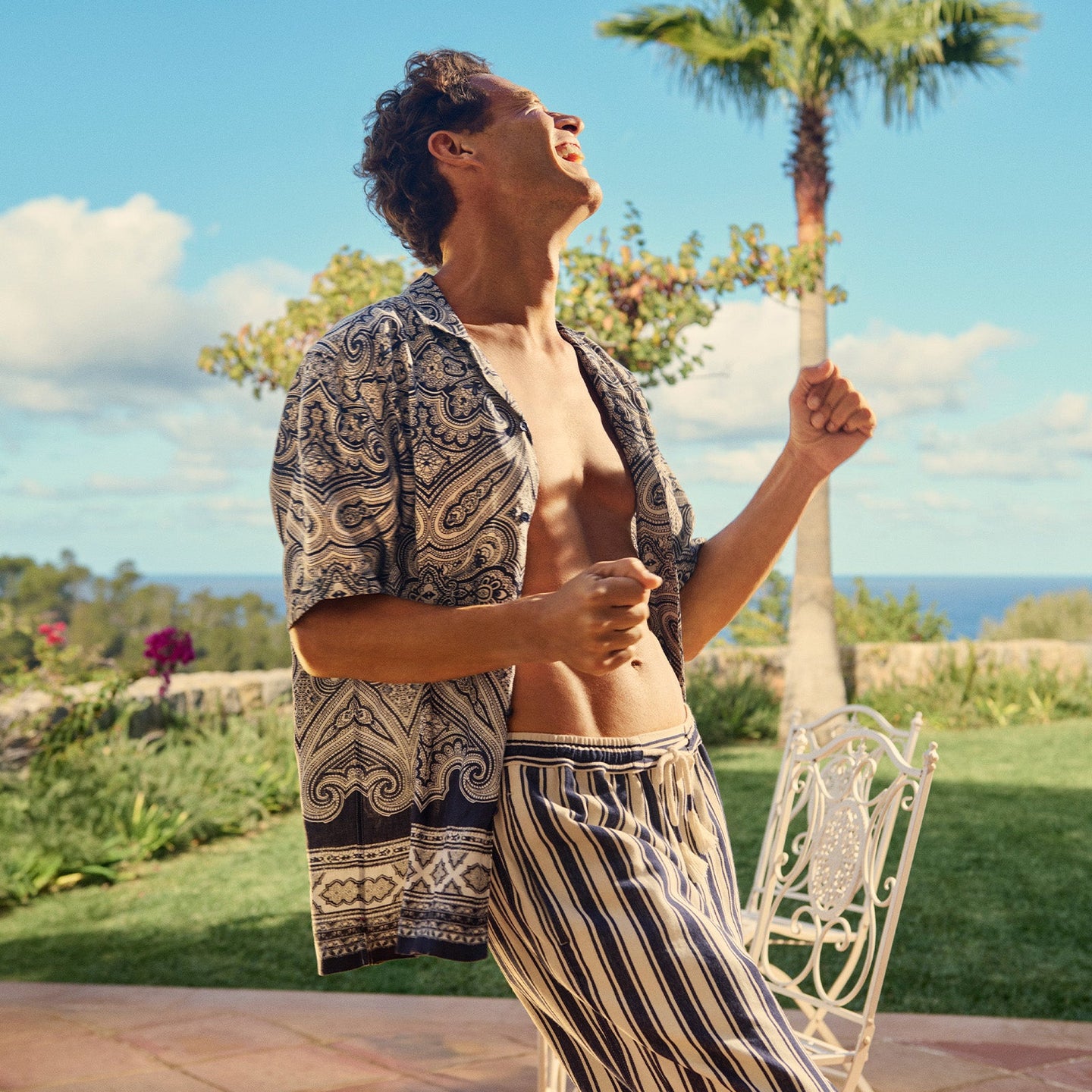 Man in patterned shirt and striped pants standing outdoors with palm trees and ocean view