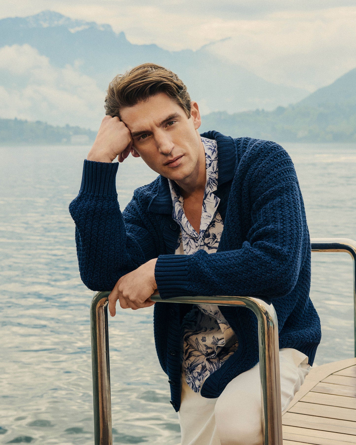 Man in a blue cardigan sitting by a lake with mountains in the background