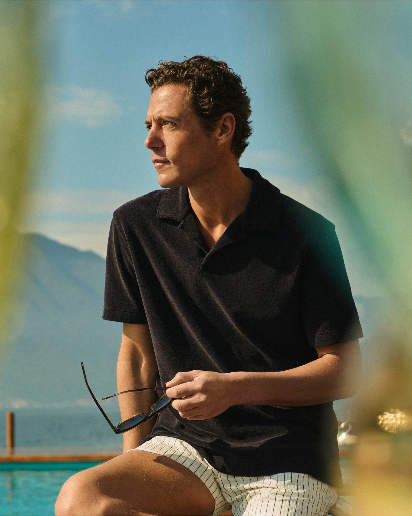 Man in a black polo shirt and white shorts sitting by a pool with mountains in the background