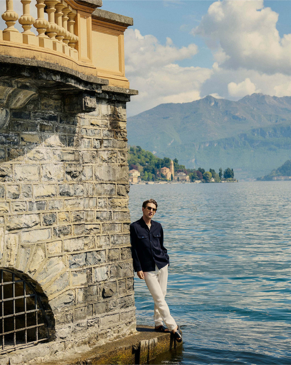 Man standing by a stone wall by a lake with mountains in the background