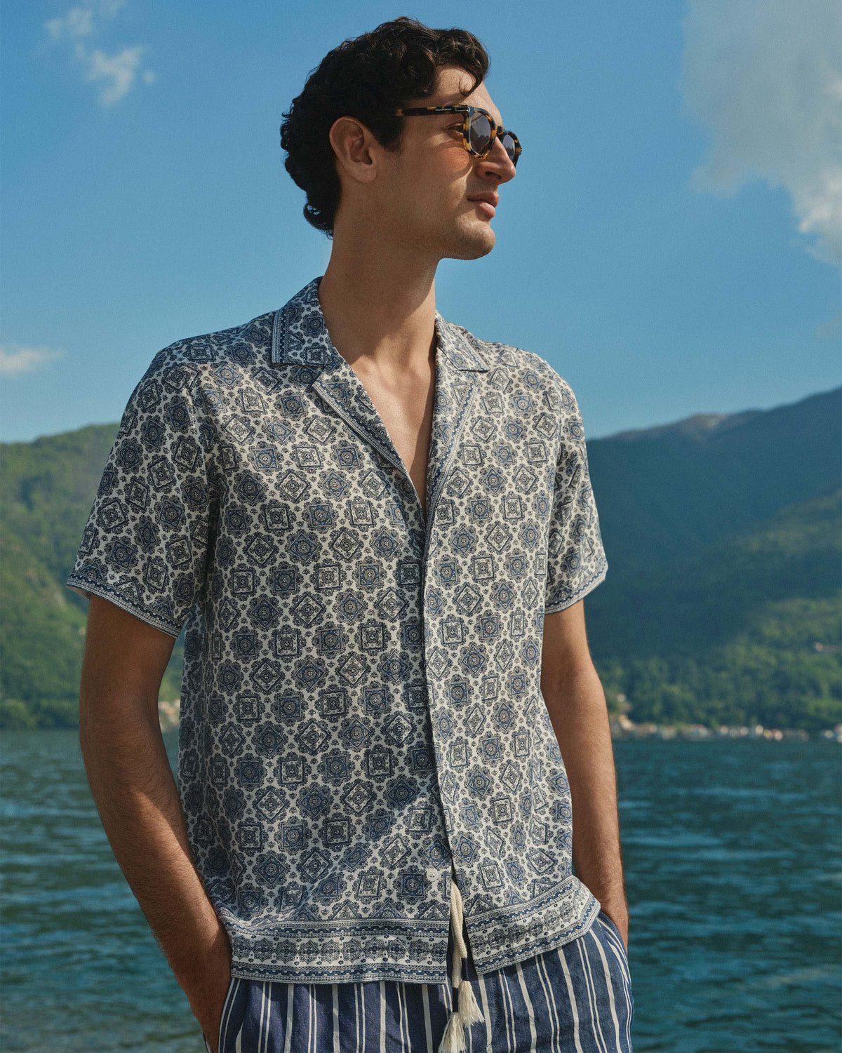 A man wearing a short-sleeve button-up shirt with a blue and white geometric pattern, standing outdoors by a lake with mountains in the background.