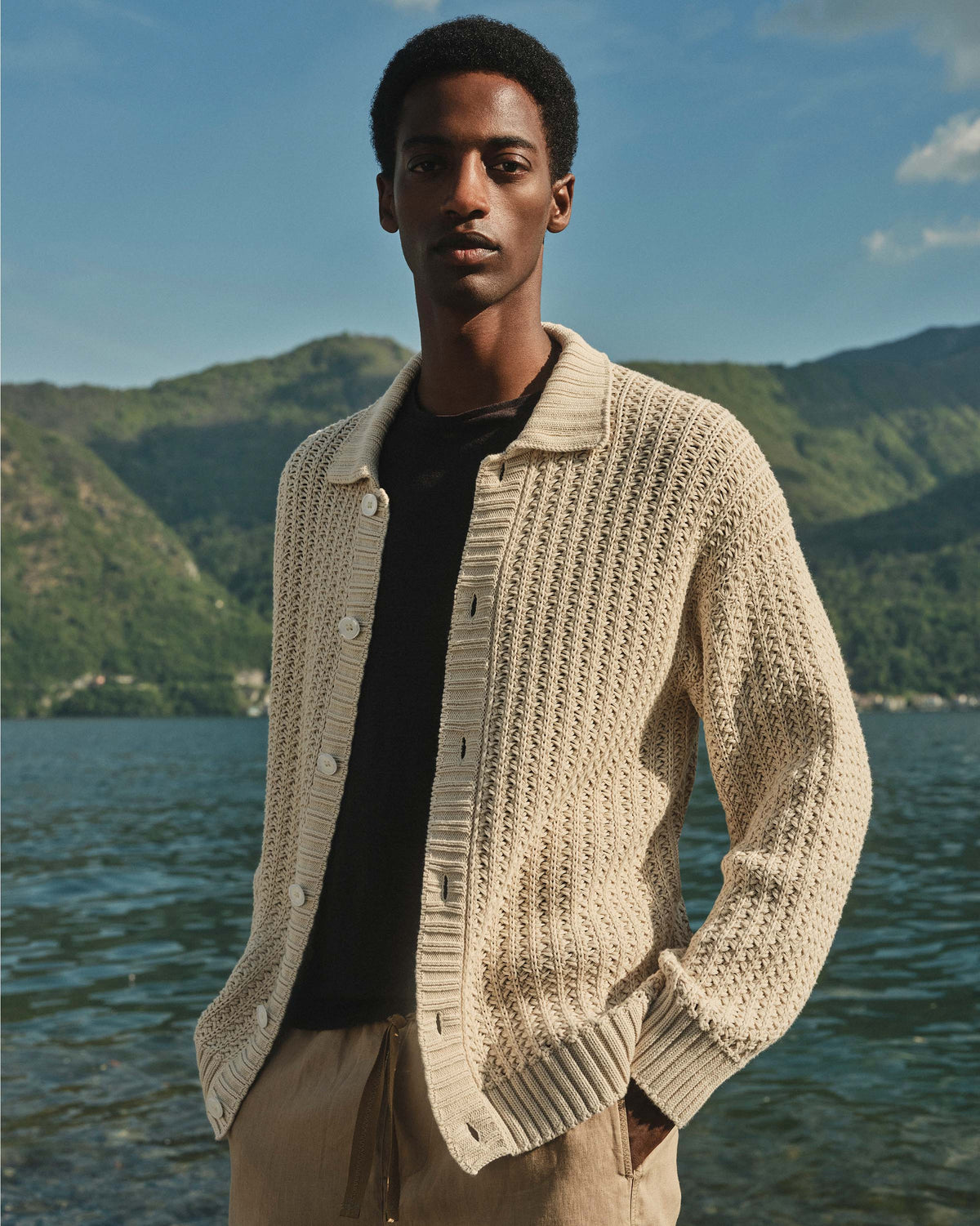 Man wearing a beige knitted cardigan standing by a lake with mountains in the background