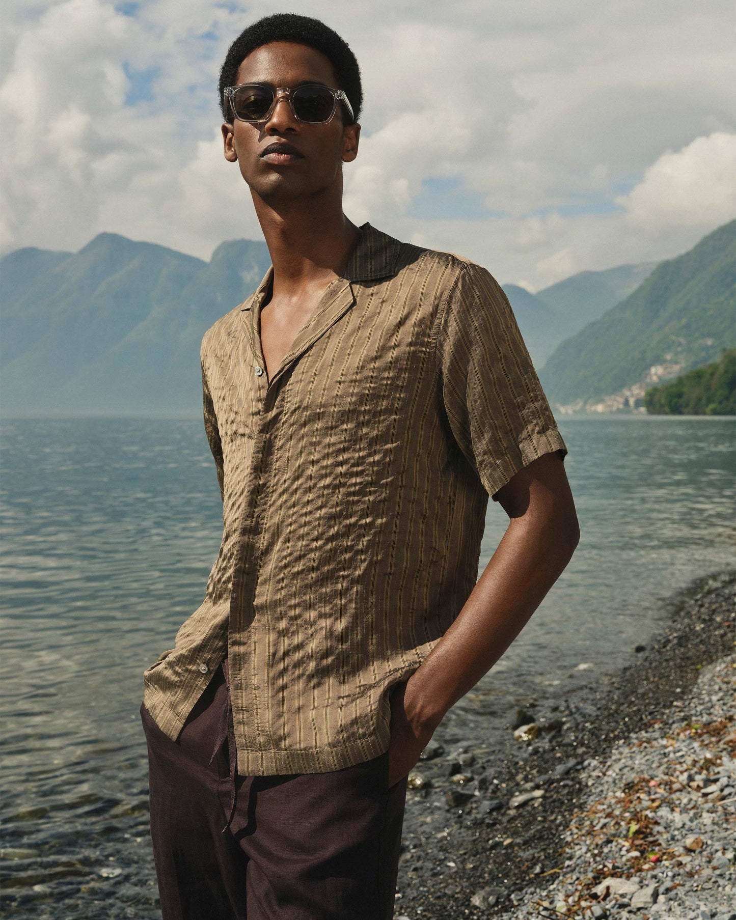 Man wearing sunglasses and a beige shirt by a lake with mountains in the background