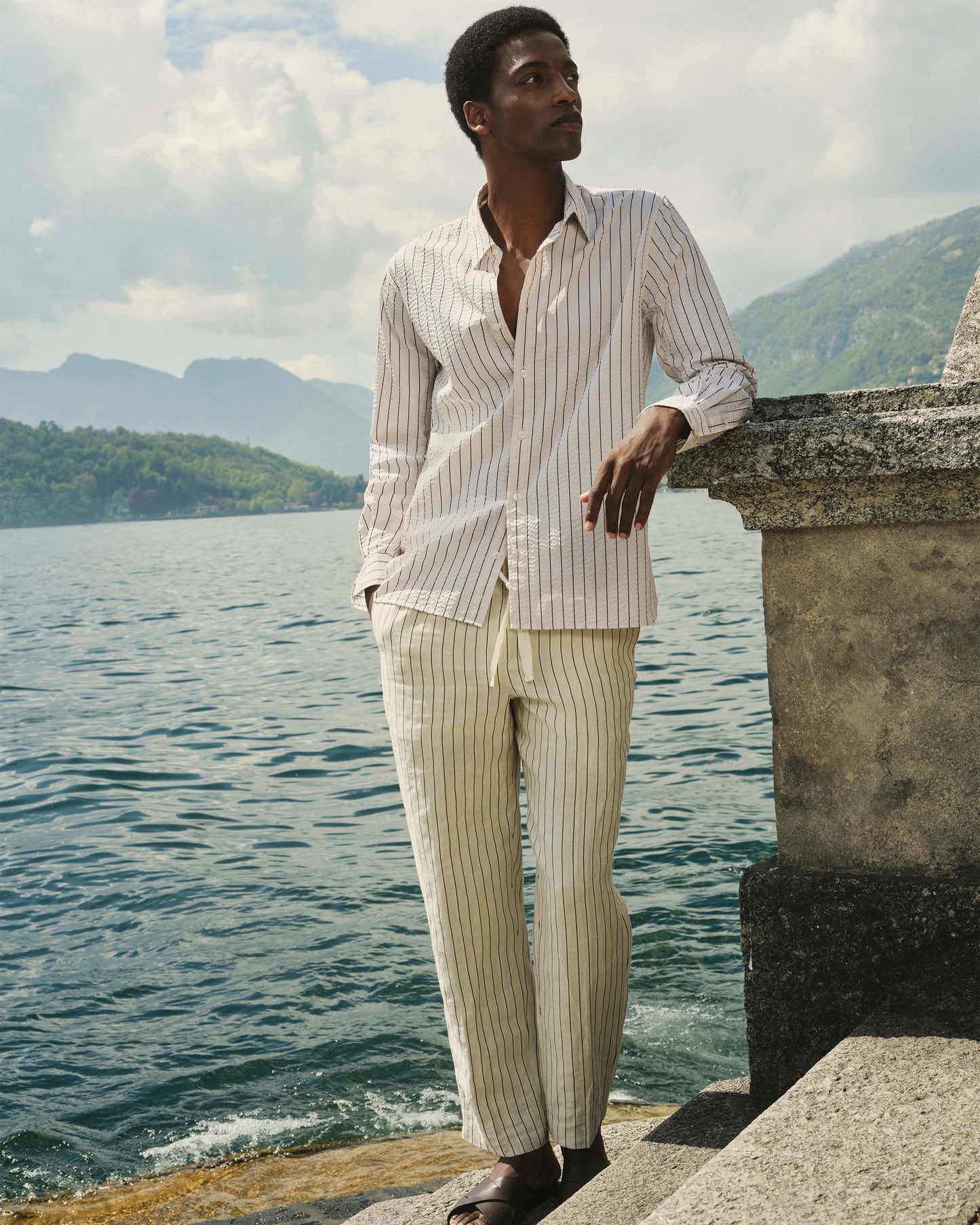 Man in striped shirt and pants standing by a body of water with mountains in the background