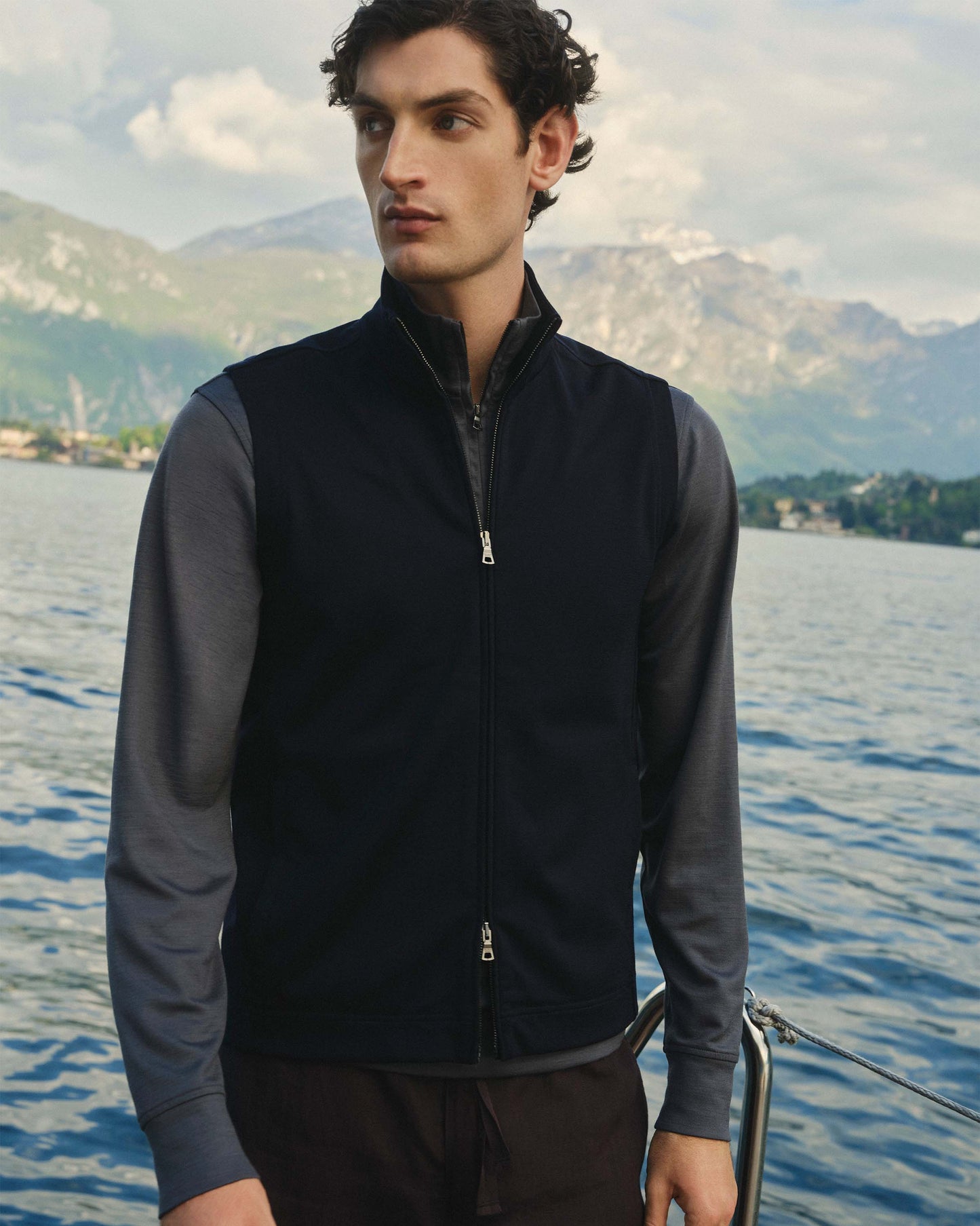 Man wearing a dark jacket standing by a body of water with mountains in the background
