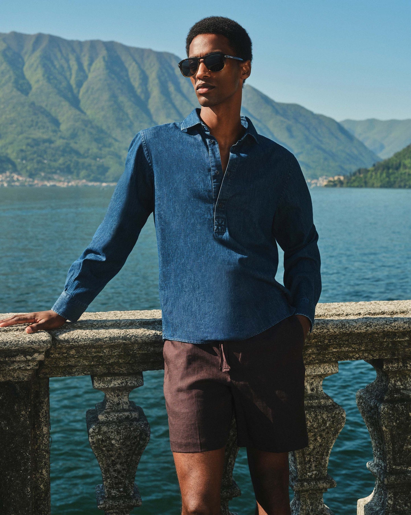 Man wearing sunglasses and a blue shirt standing by a scenic lakeside with mountains in the background.