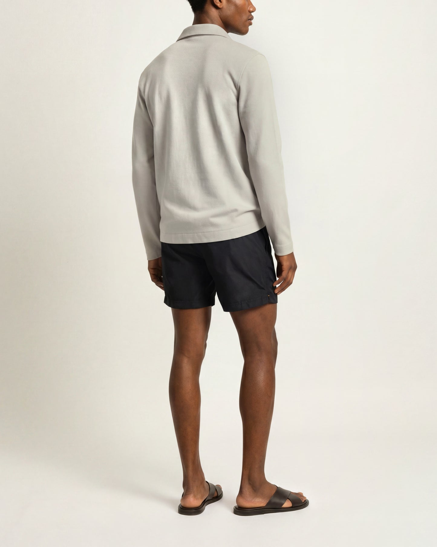 A man shown from the back wearing a light gray long-sleeve polo shirt, dark navy shorts, and dark brown slide sandals, standing against a plain off-white studio background.