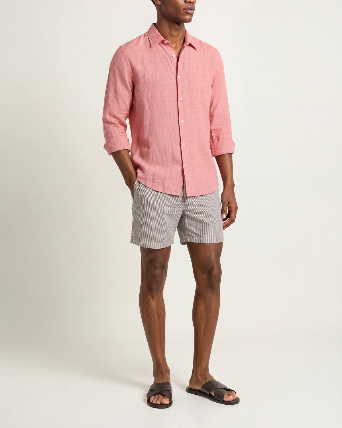 A man modeling a light pink linen button-down shirt with rolled-up sleeves, paired with gray shorts and black slide sandals, against a plain off-white studio background.