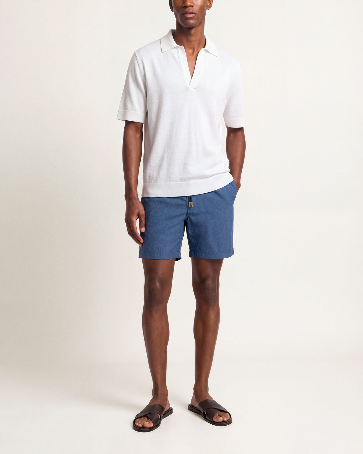 A man modeling a white short-sleeve polo shirt and blue shorts with dark brown slide sandals, standing against a plain off-white studio background.