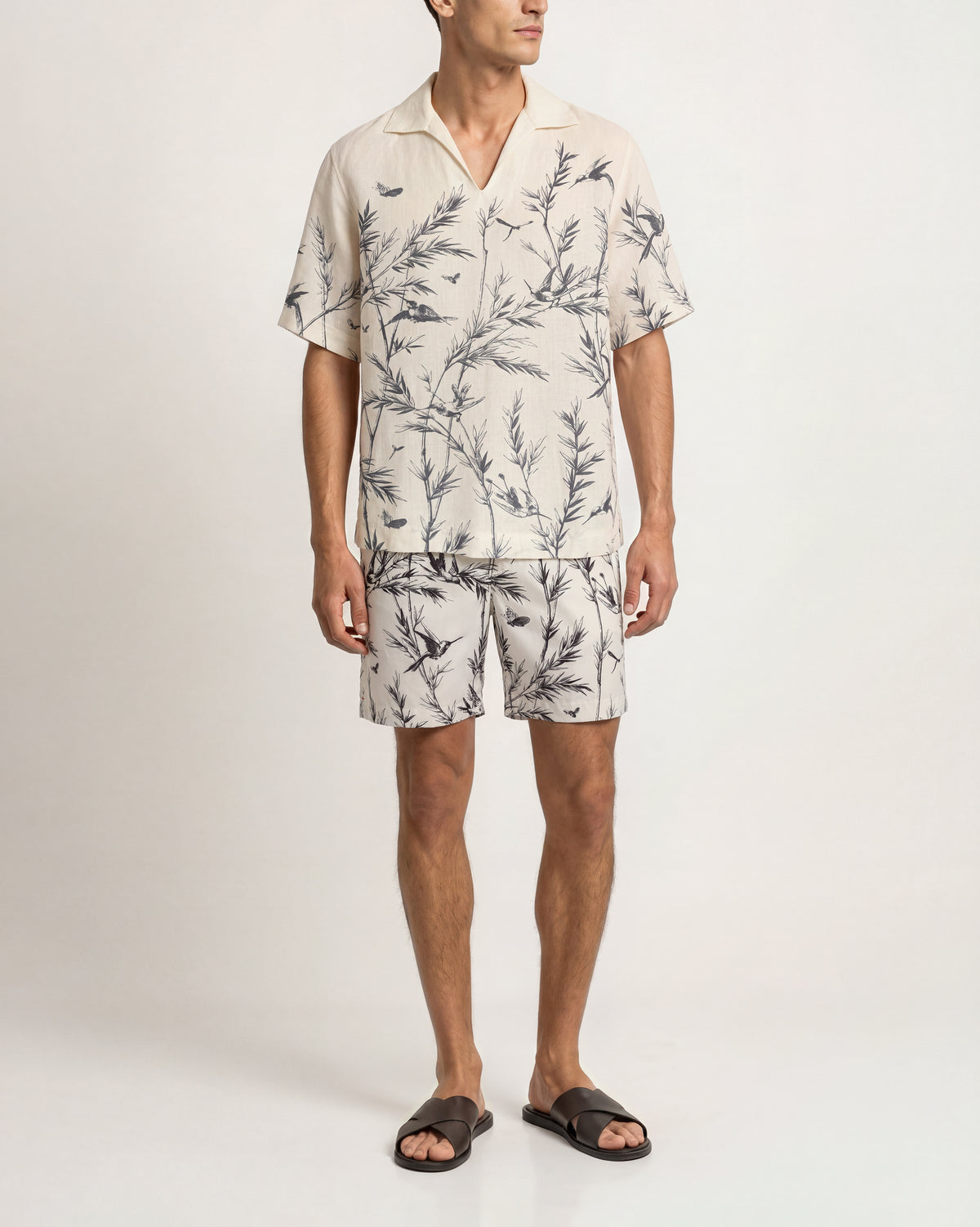 A man modeling a matching short-sleeve camp collar shirt and shorts set in a light beige or off-white fabric with a dark gray botanical leaf print, paired with dark brown slide sandals, against a plain light gray studio background.