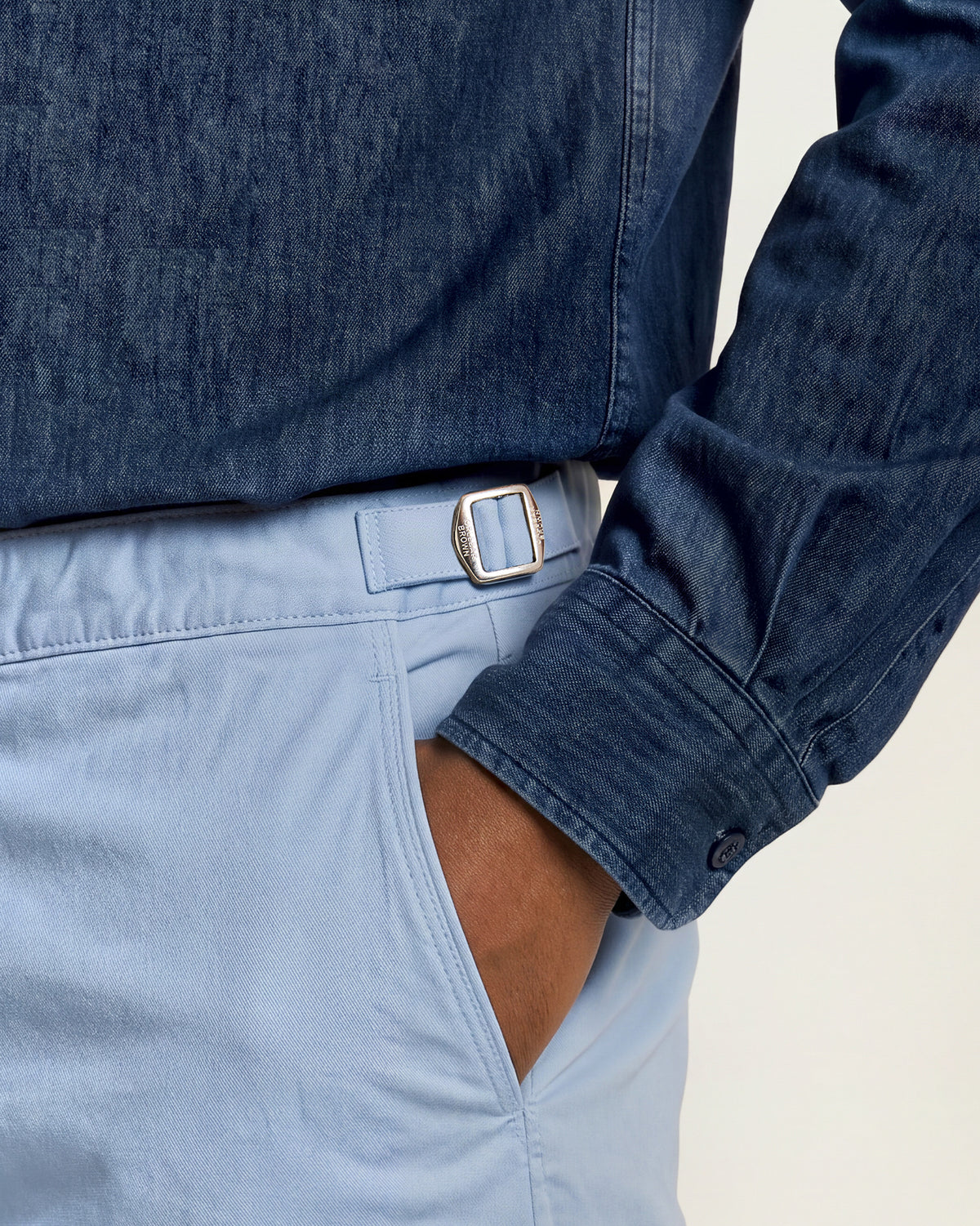 Closeup of blue trousers with silver side adjuster, worn with a dark denim shirt on a plain background