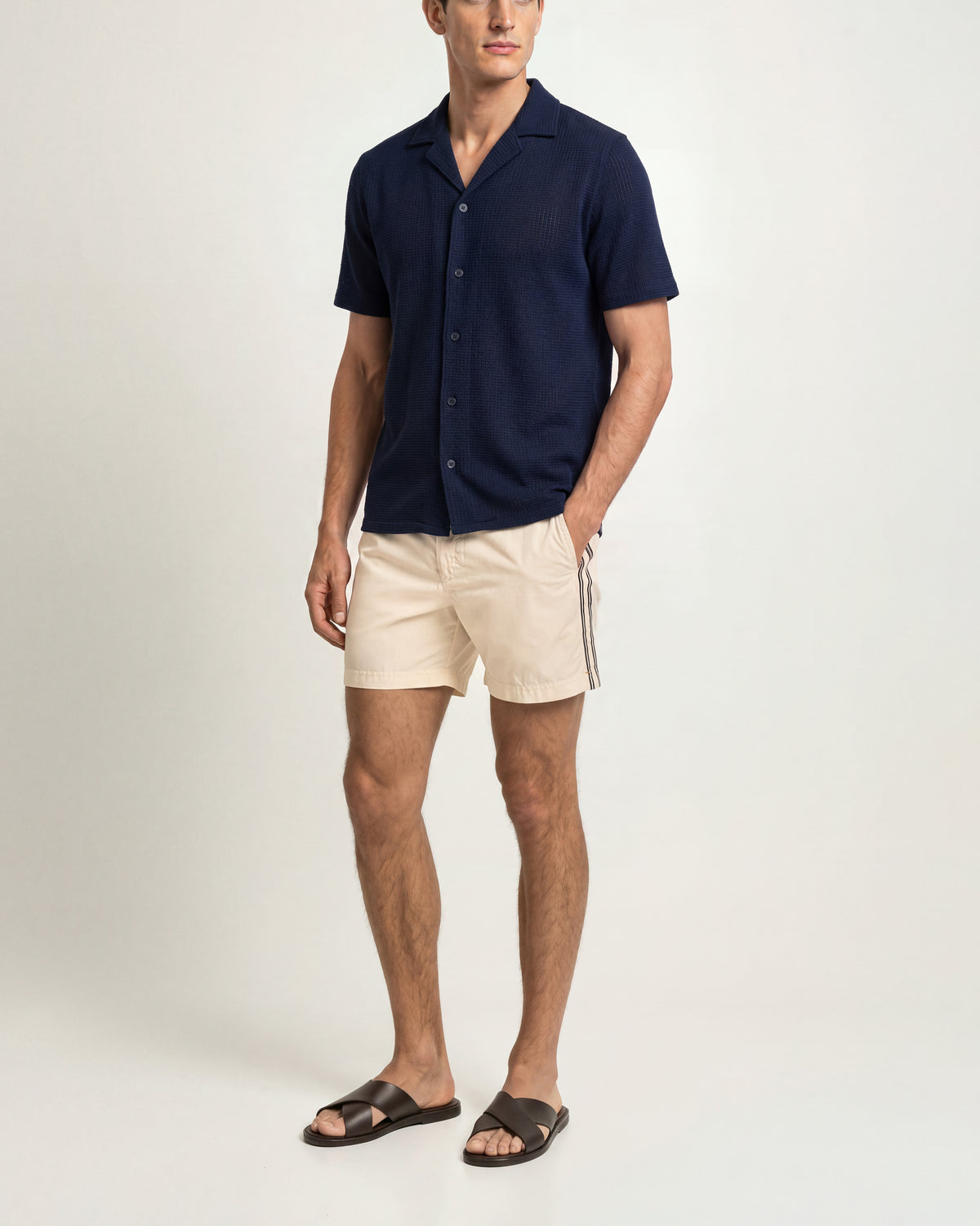 A man modeling a short-sleeve navy blue button-up shirt and beige shorts with white side stripes, paired with dark brown slide sandals, against a light gray studio background.