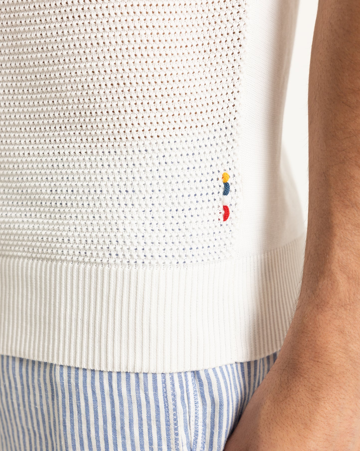 Close-up of a white textured sweater with a small embroidered logo, worn over a blue and white striped shirt.