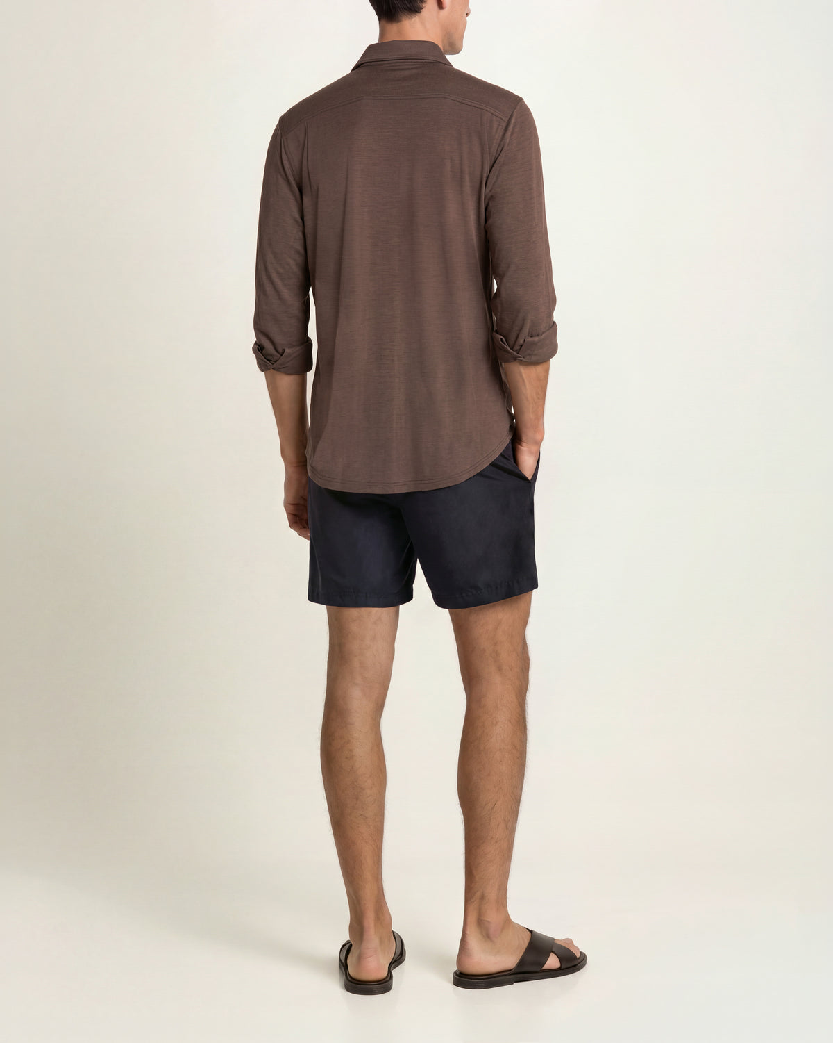 Man wearing brown shirt with black shorts and brown sandals on a plain background