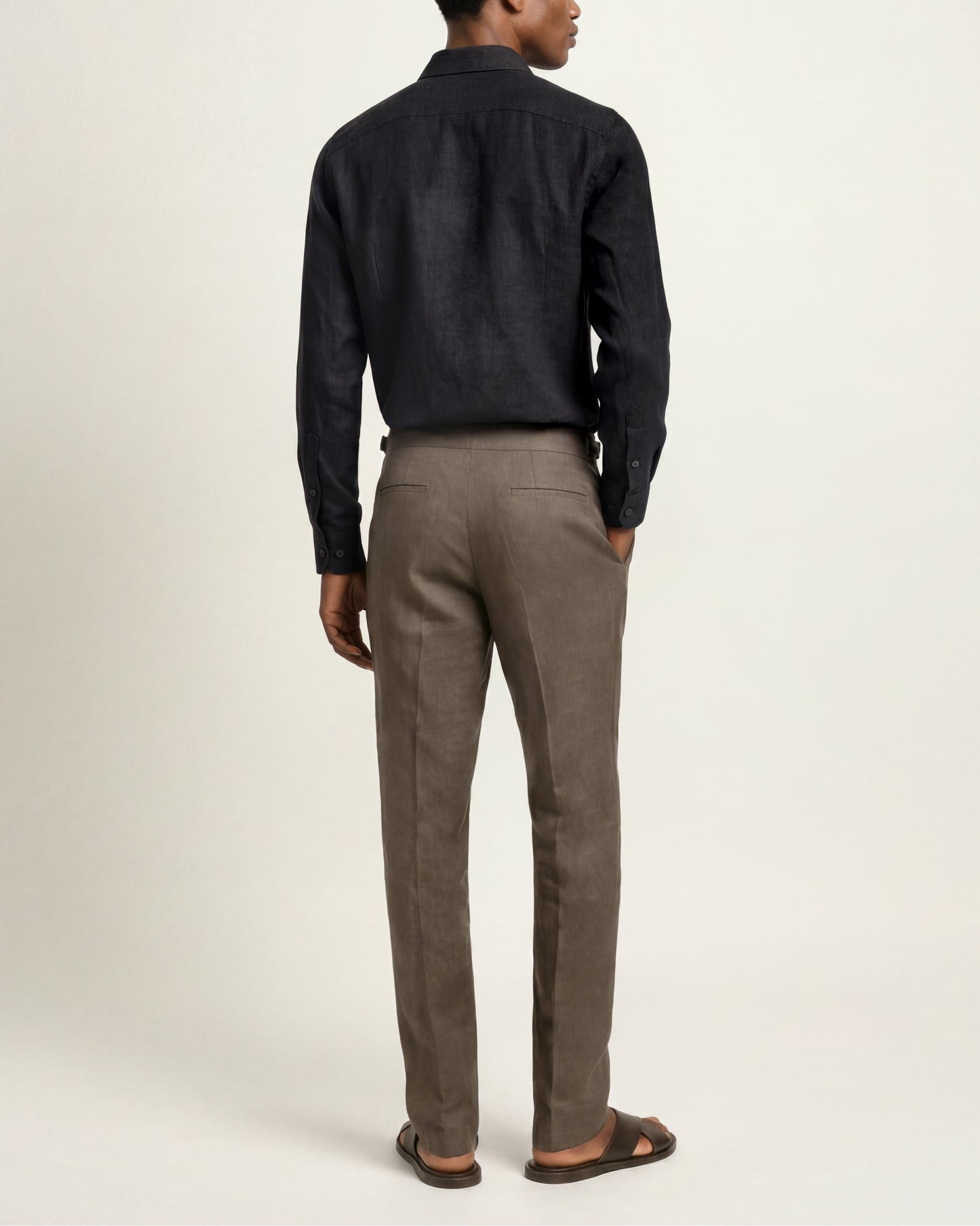 A man shown from the back wearing a dark charcoal long-sleeve shirt and brown trousers, standing against a plain off-white studio background.