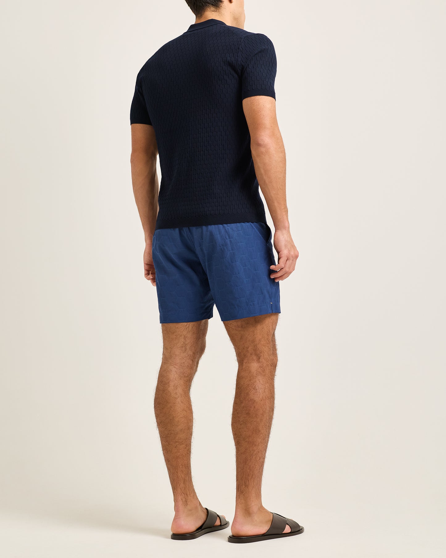 Man wearing navy textured polo shirt with navy swim shorts on a plain background