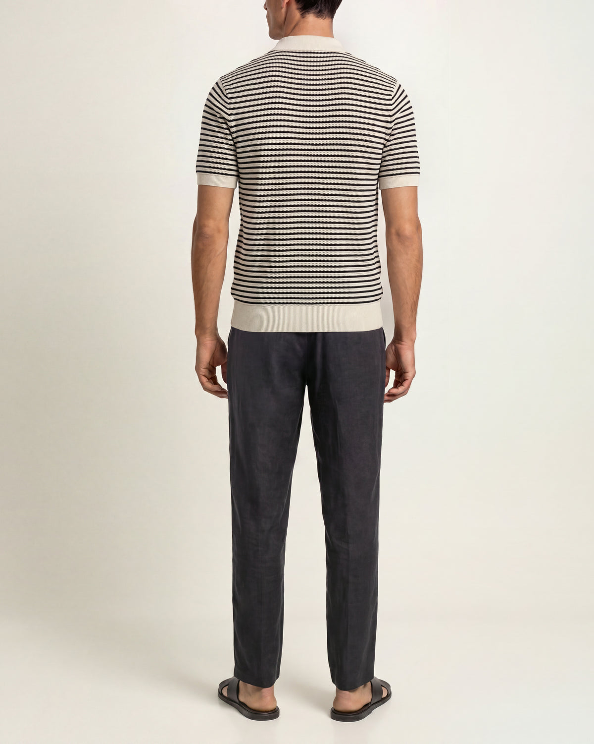A man viewed from the back wearing a short-sleeve knit top with thin horizontal black and beige stripes, paired with dark charcoal trousers and black sandals, against a plain off-white studio background.