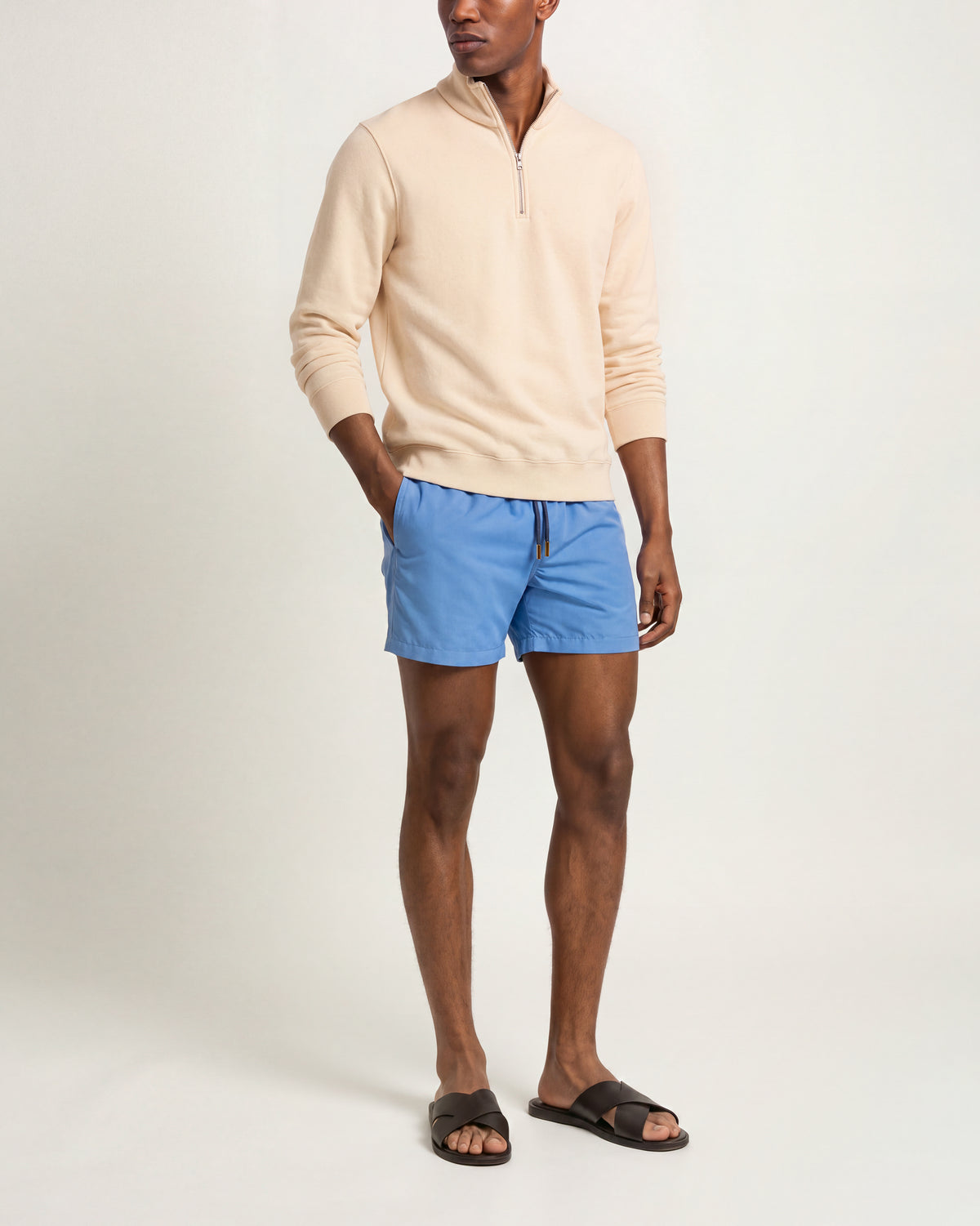 A man modeling a light beige quarter-zip long-sleeve sweater, blue drawstring shorts, and black slide sandals against a plain light gray studio background.