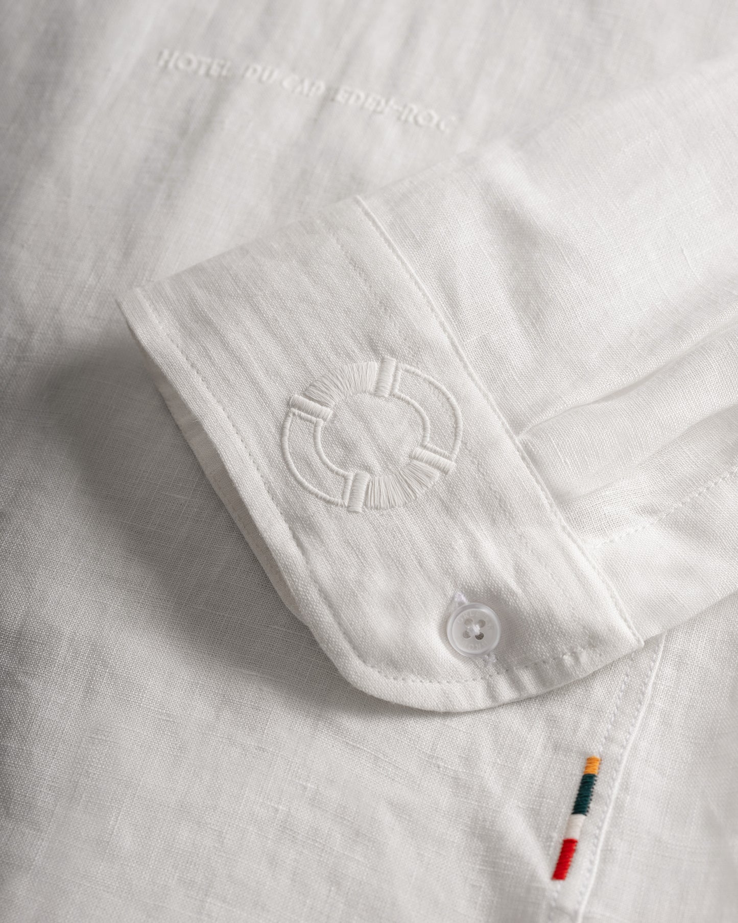 Close-up of a white linen shirt sleeve with a buttoned cuff, tonal embroidered logo, and a small colorful striped accent tab near the hem.