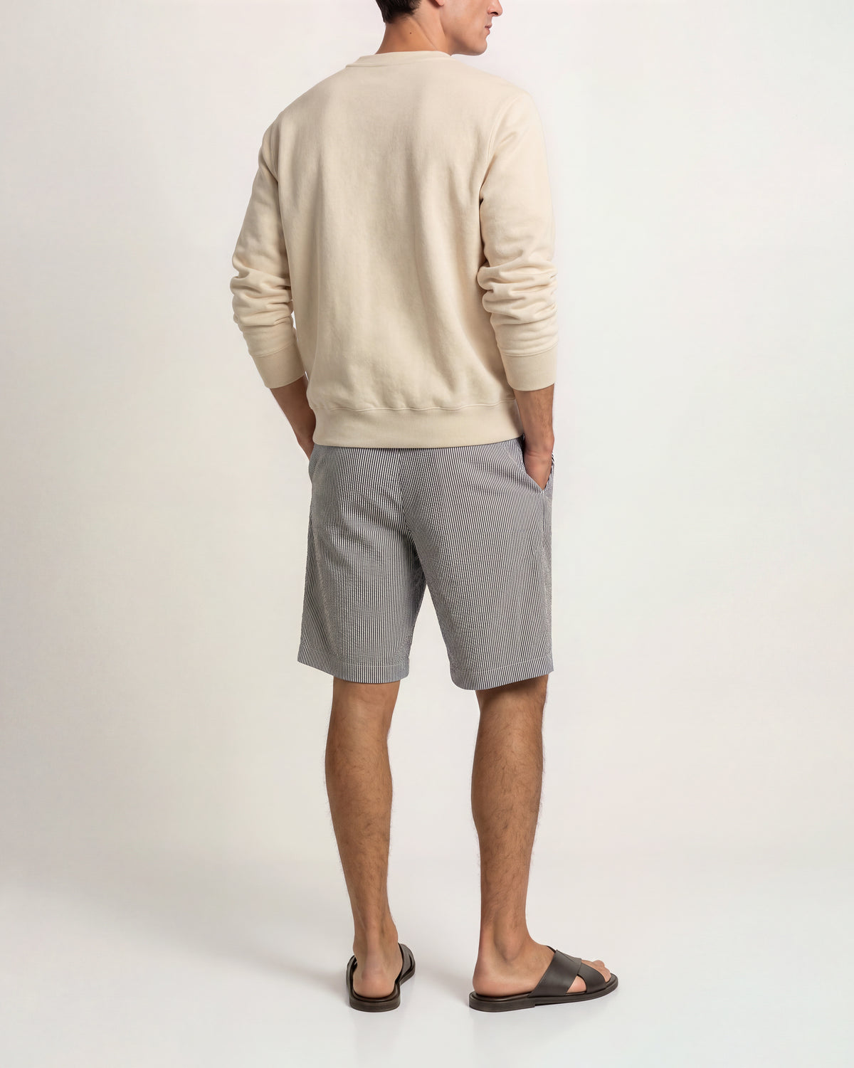 A man is shown from behind wearing a cream-colored long-sleeve crewneck sweatshirt, light gray linen shorts, and dark brown slide sandals against a plain white studio background.