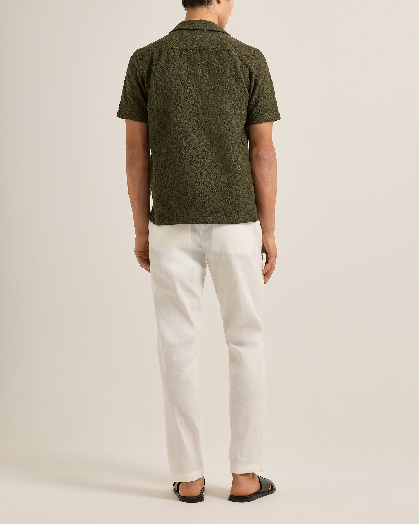 Person wearing a green patterned shirt and white pants on a beige background