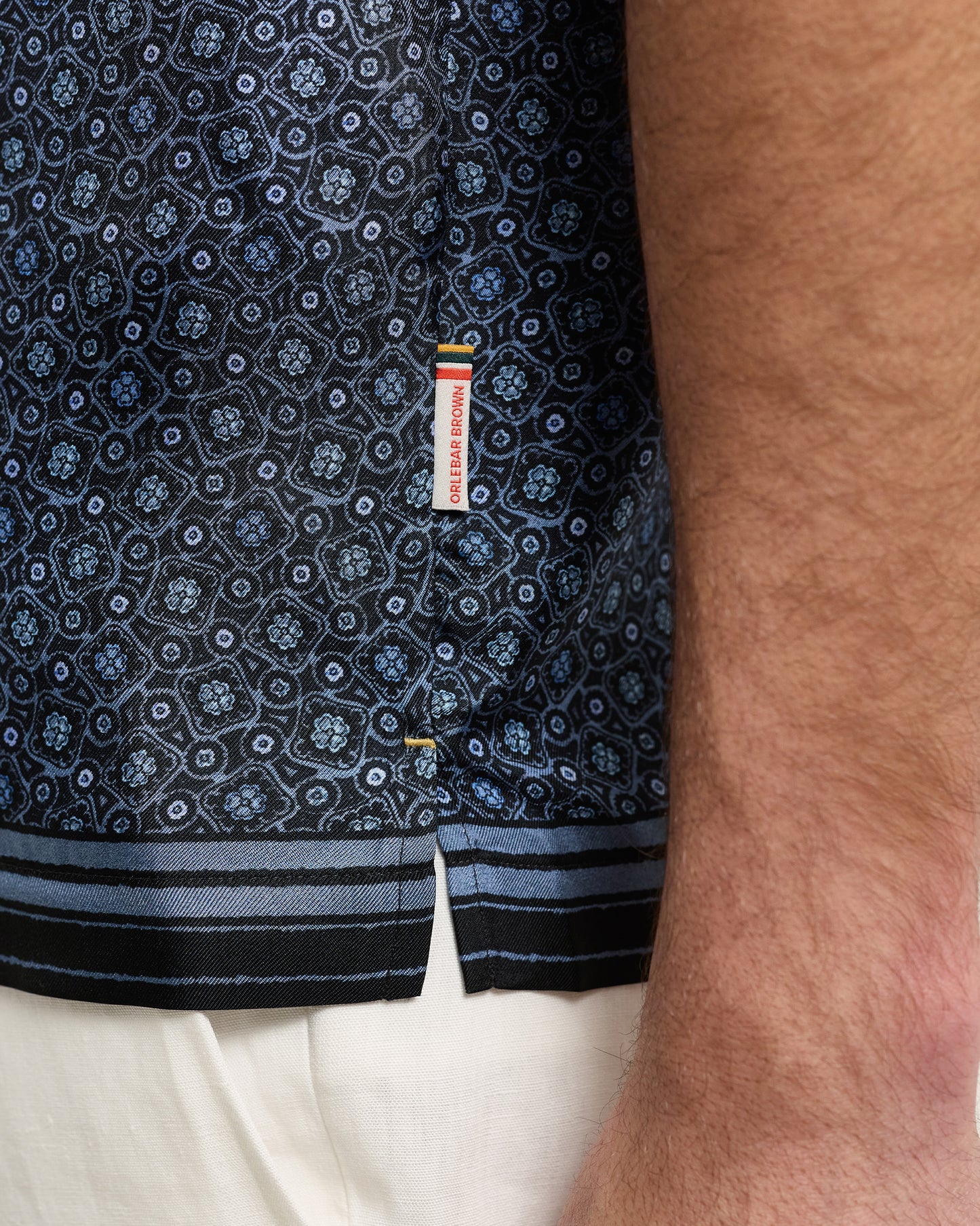 Close-up of a patterned shirt with a brand label on a person wearing white pants.