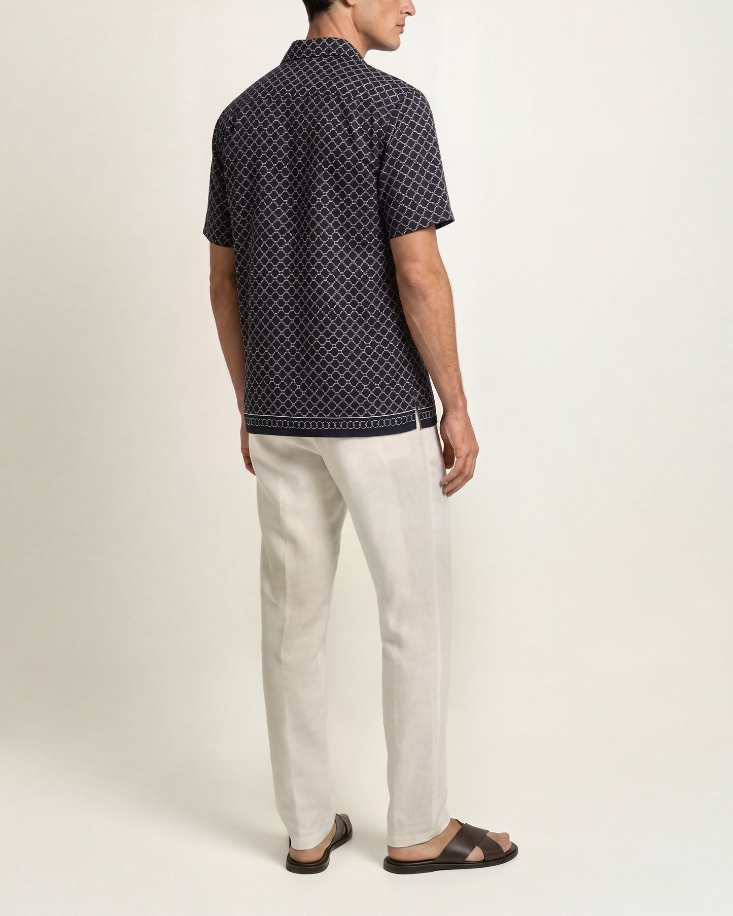 A man shown from the back wearing a short-sleeve button-up shirt with a small white geometric pattern on a dark navy background, paired with off-white trousers and dark brown sandals, against a plain light beige studio background.