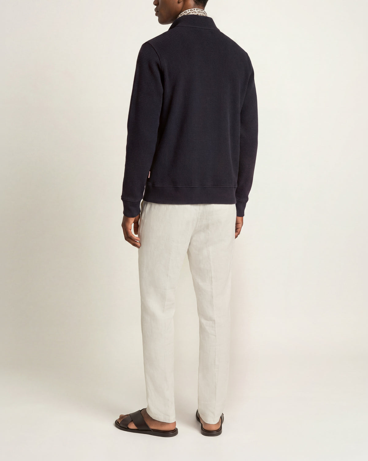 Man wearing navy long sleeve sweatshirt with white trousers and brown sandals on a plain background