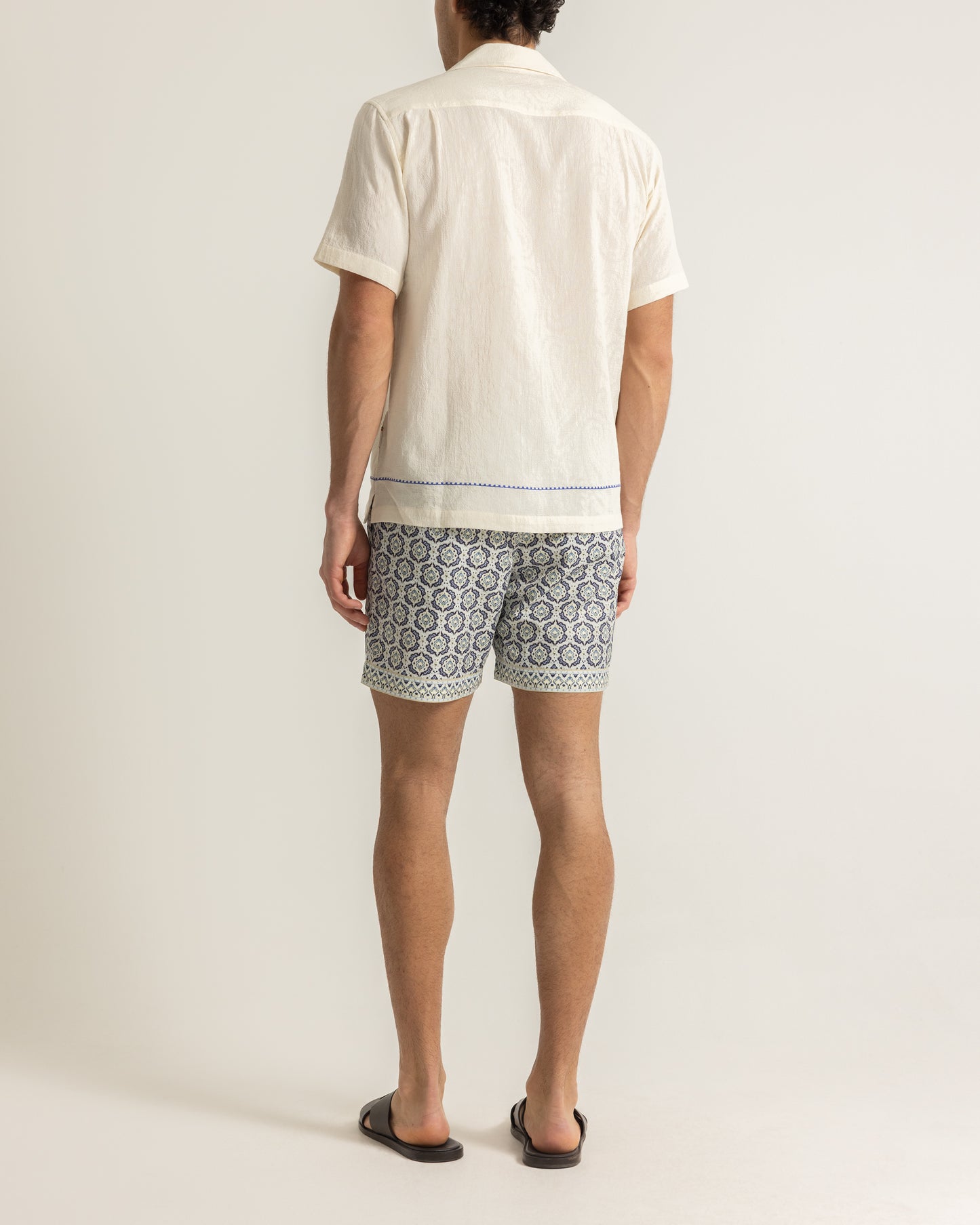 Man wearing white printed white shirt with patterned swim shorts on a plain background