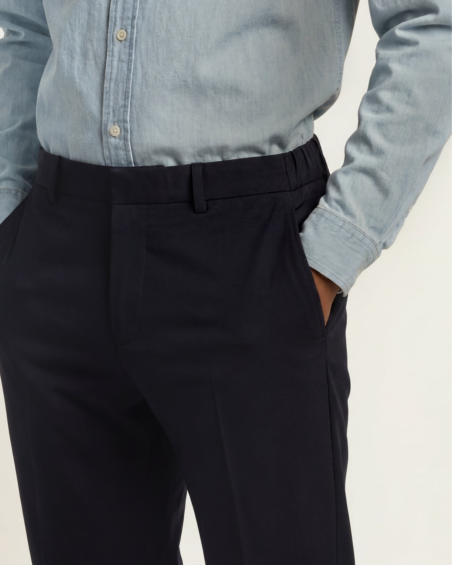Close-up of a person wearing dark navy trousers and a light blue long-sleeve button-up shirt, with one hand in a trouser pocket, against a plain light background.