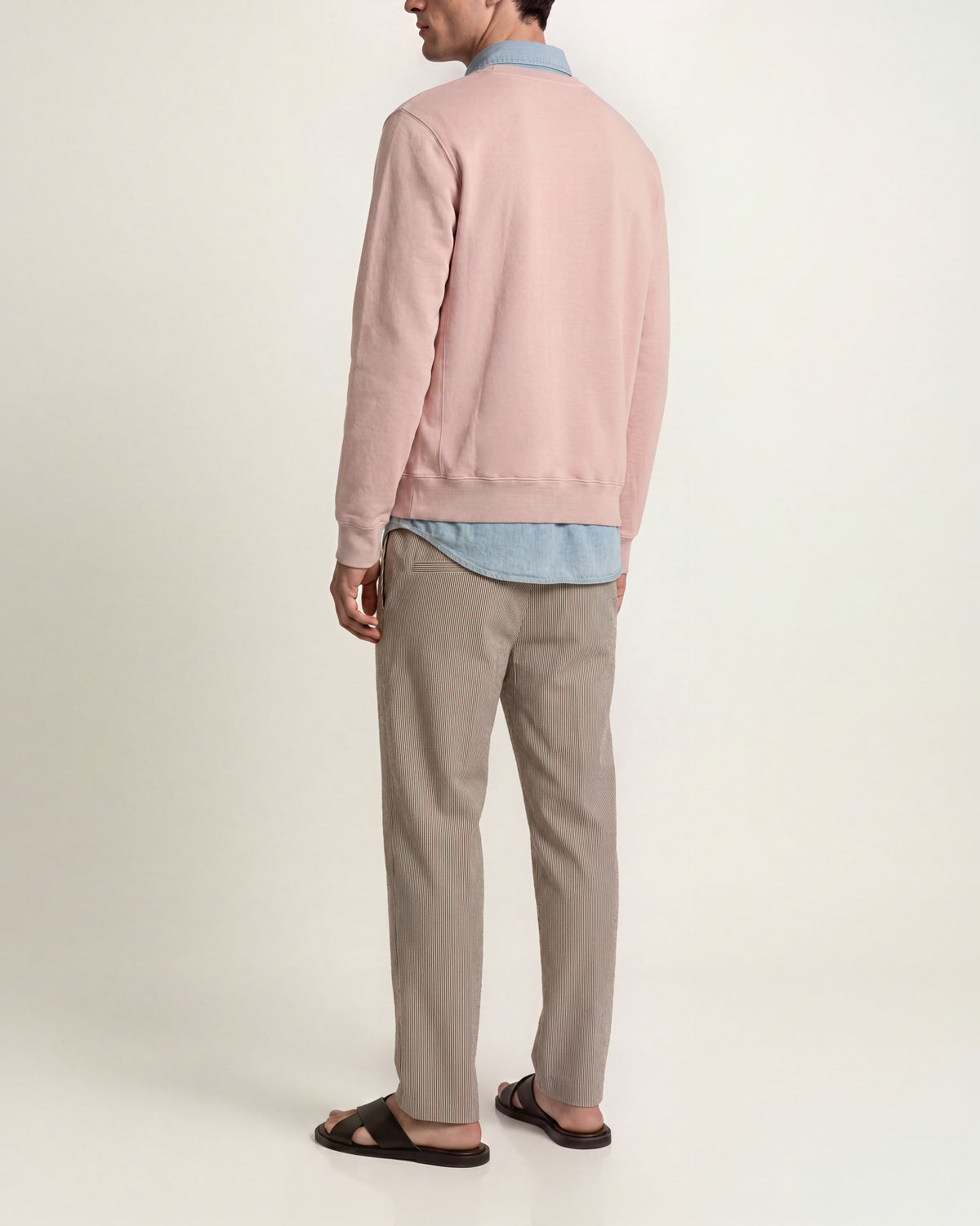 Man wearing pink sweatshirt with denim shirt underneath and brown/ white striped trousers on a plain background