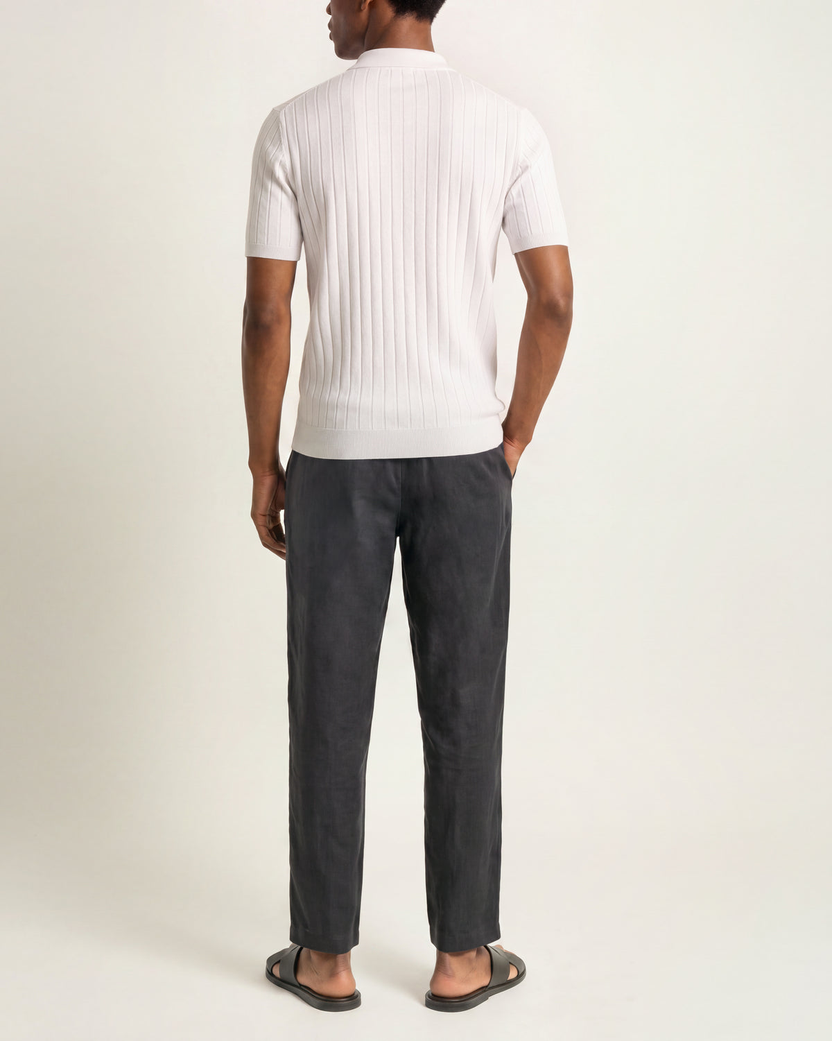 A man shown from the back wearing a white ribbed short-sleeve knit top, dark charcoal gray trousers, and black slide sandals, photographed against a plain light studio background.