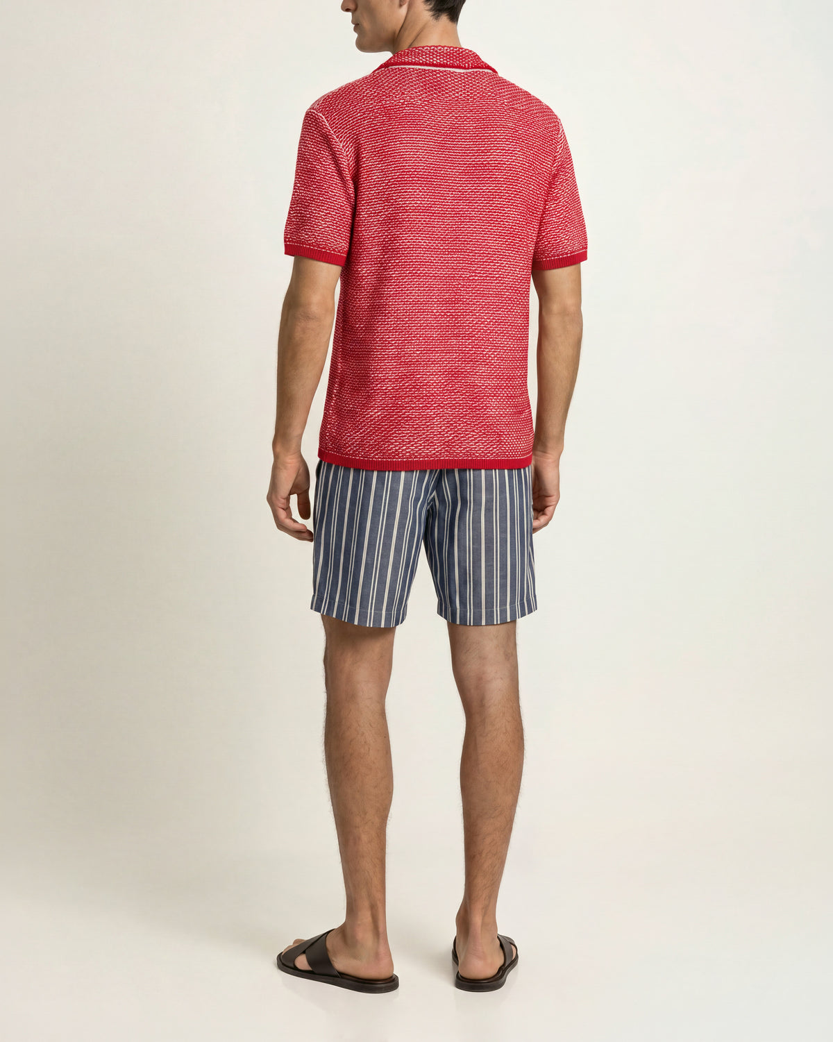 Back view of a man wearing a red-and-white striped short-sleeved crochet polo shirt, blue striped drawcord-waist shorts, and brown sandals on a plain background