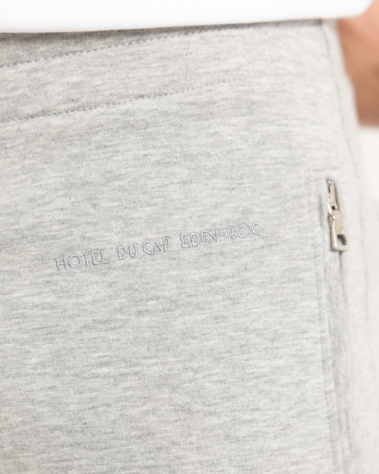 Close-up of light heather gray sweatpants showing the side pocket with a silver zipper and the embroidered text 'HOTEL DU CAP EDEN-ROC' on the front.