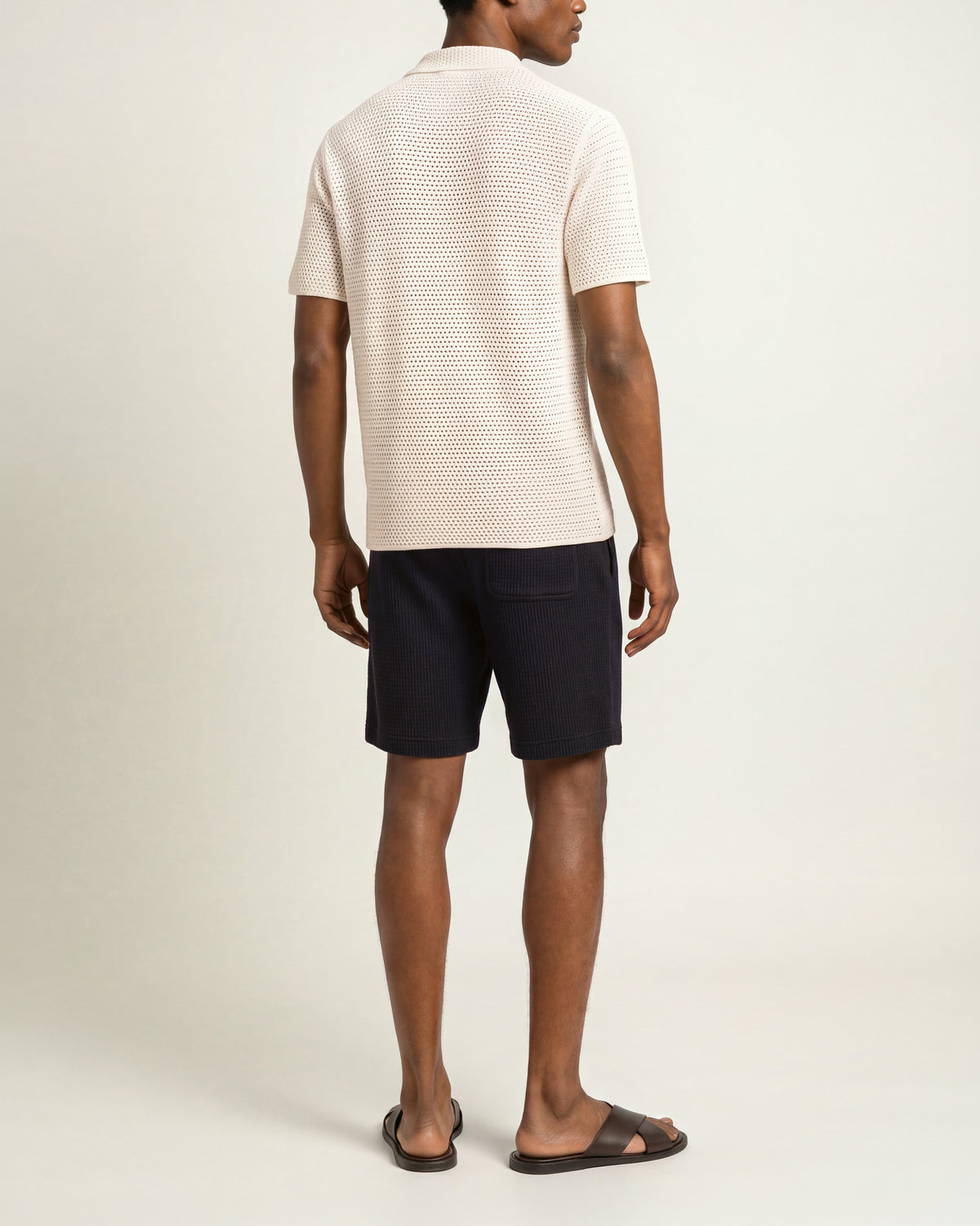 Man wearing white crochet stich polo shirt with navy towelling shorts on a plain background