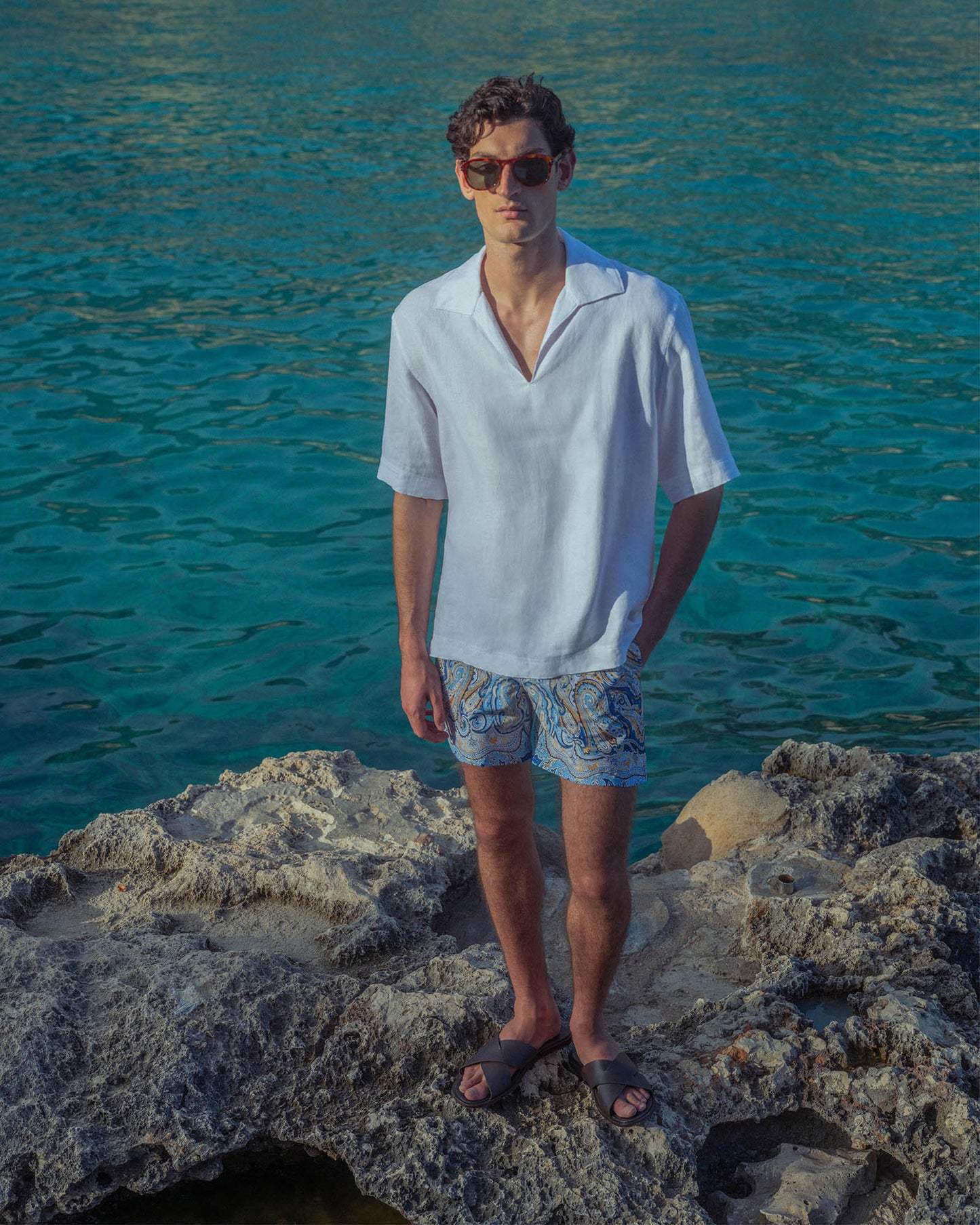 Man standing on rocks by the ocean wearing a white shirt and patterned shorts.