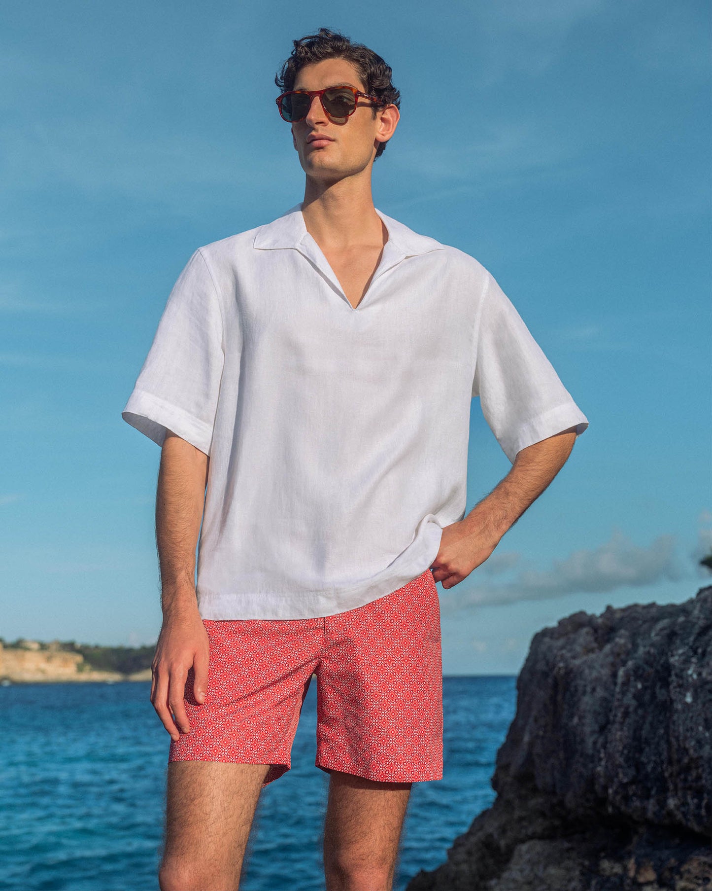 Man wearing white short sleeve shirt with red printed swim shorts, against backdrop an the ocean