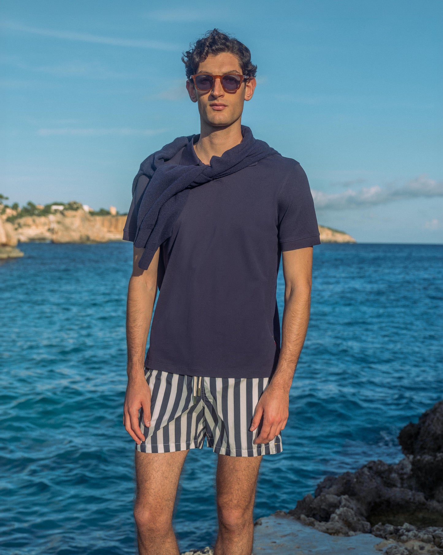 Man wearing sunglasses with navy polo shirt and striped shorts standing by the ocean