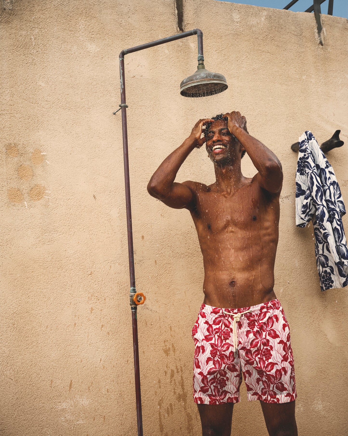 Man in red floral swim shorts standing under an outdoor shower against a beige wall.