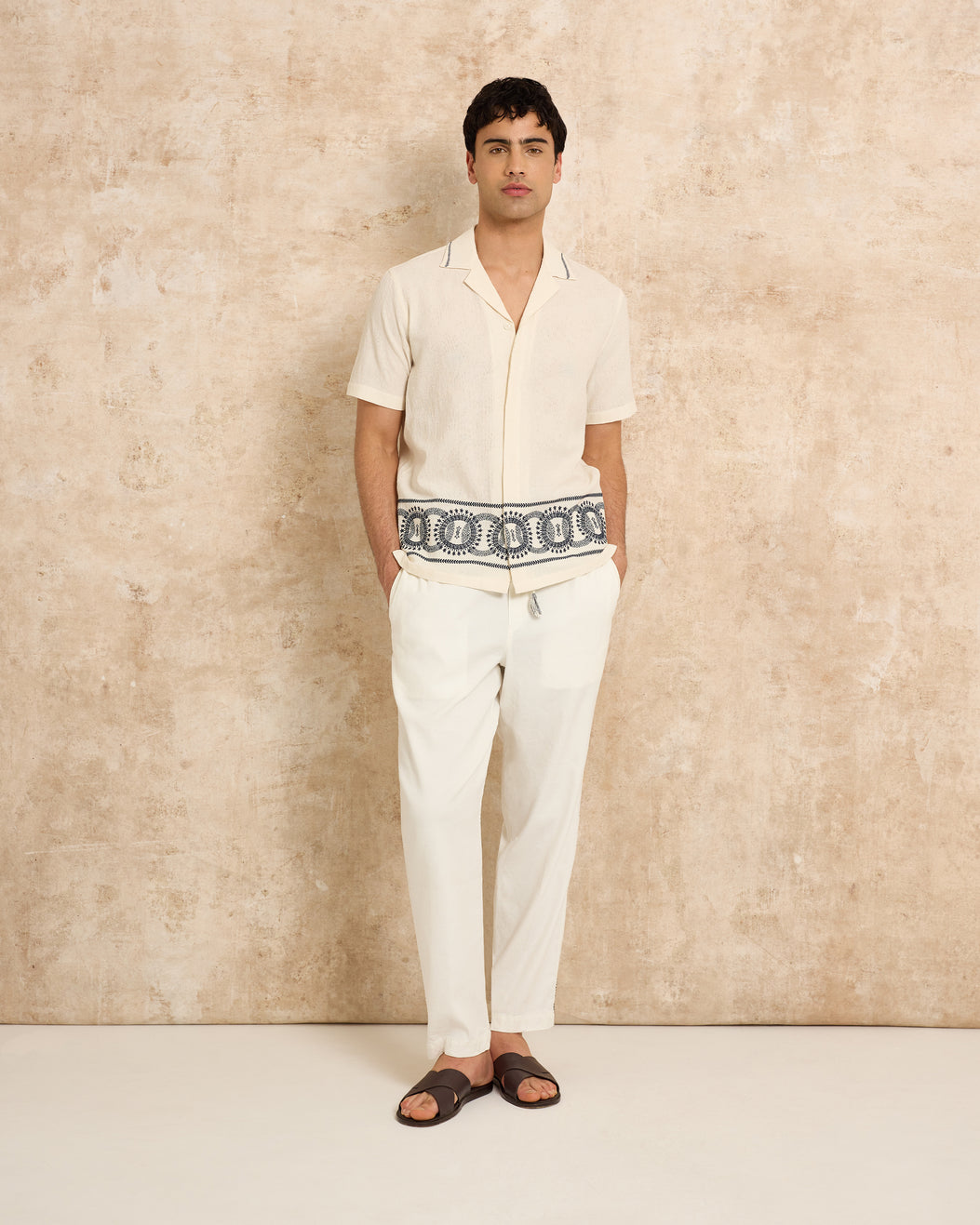 Man wearing a white shirt and pants with a patterned trim against a beige wall.
