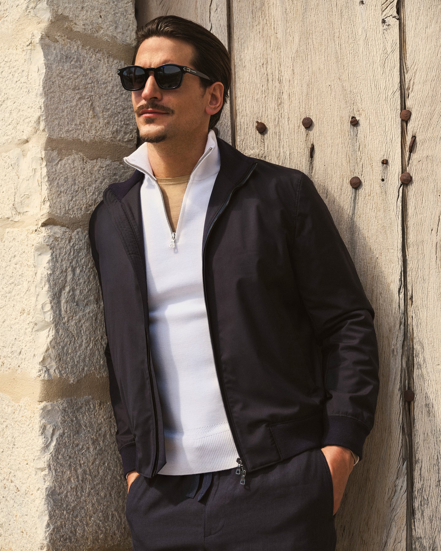 Man wearing sunglasses and a black jacket leaning against a stone wall.