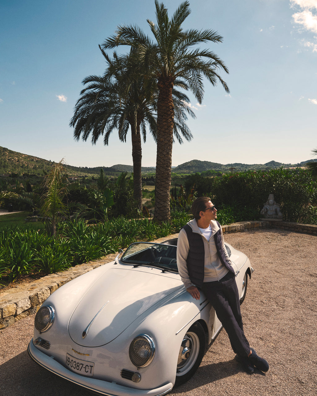 Person leaning against a vintage car with palm trees and mountains in the background