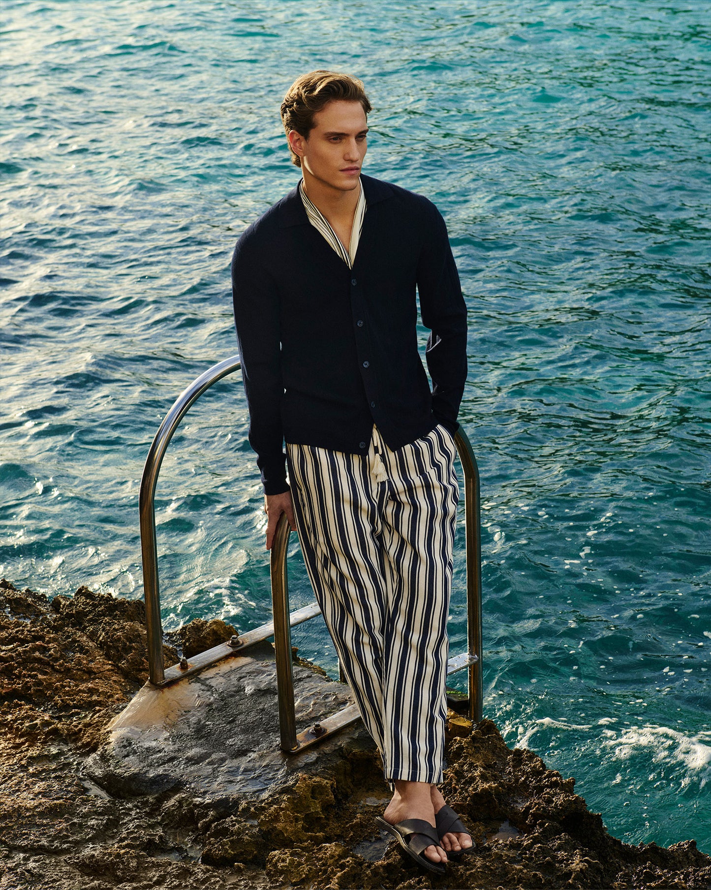 Man wearing navy knit cardigan with stripe trousers against backdrop of the ocean