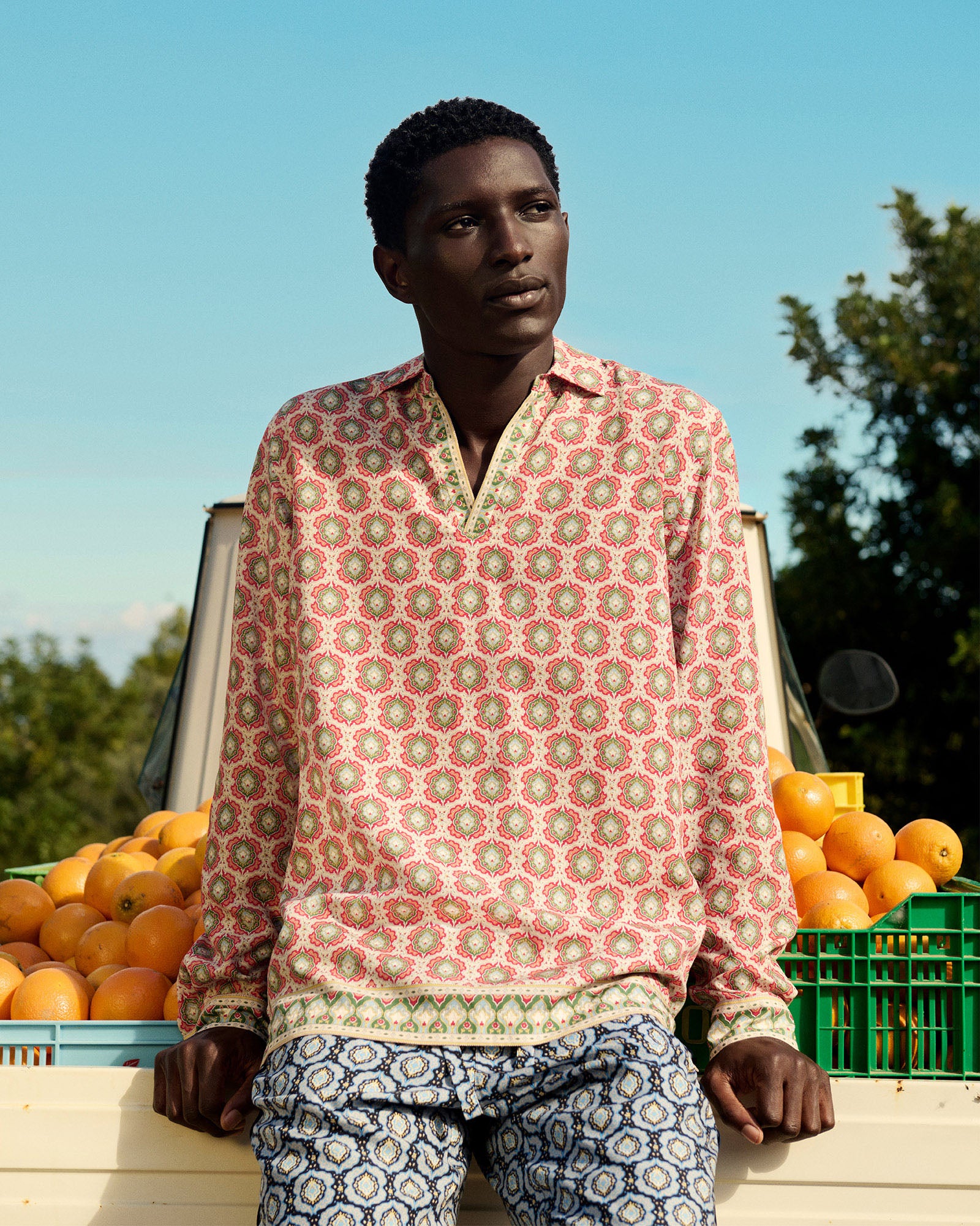Man wearing a patterned shirt and pants standing in front of stacked oranges with a clear blue sky.