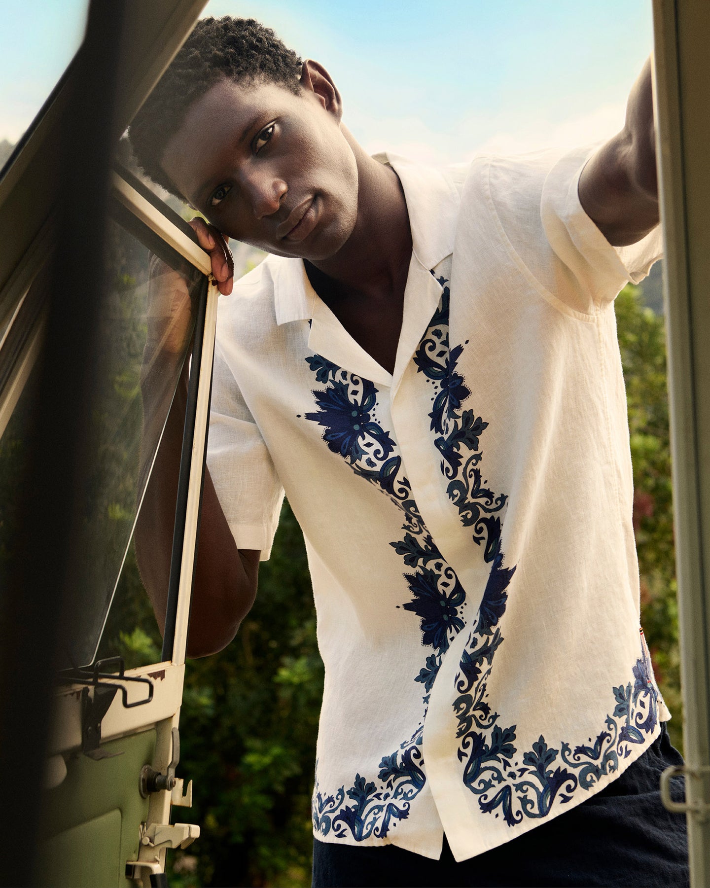 Man wearing a white shirt with blue floral patterns leaning against a vehicle.