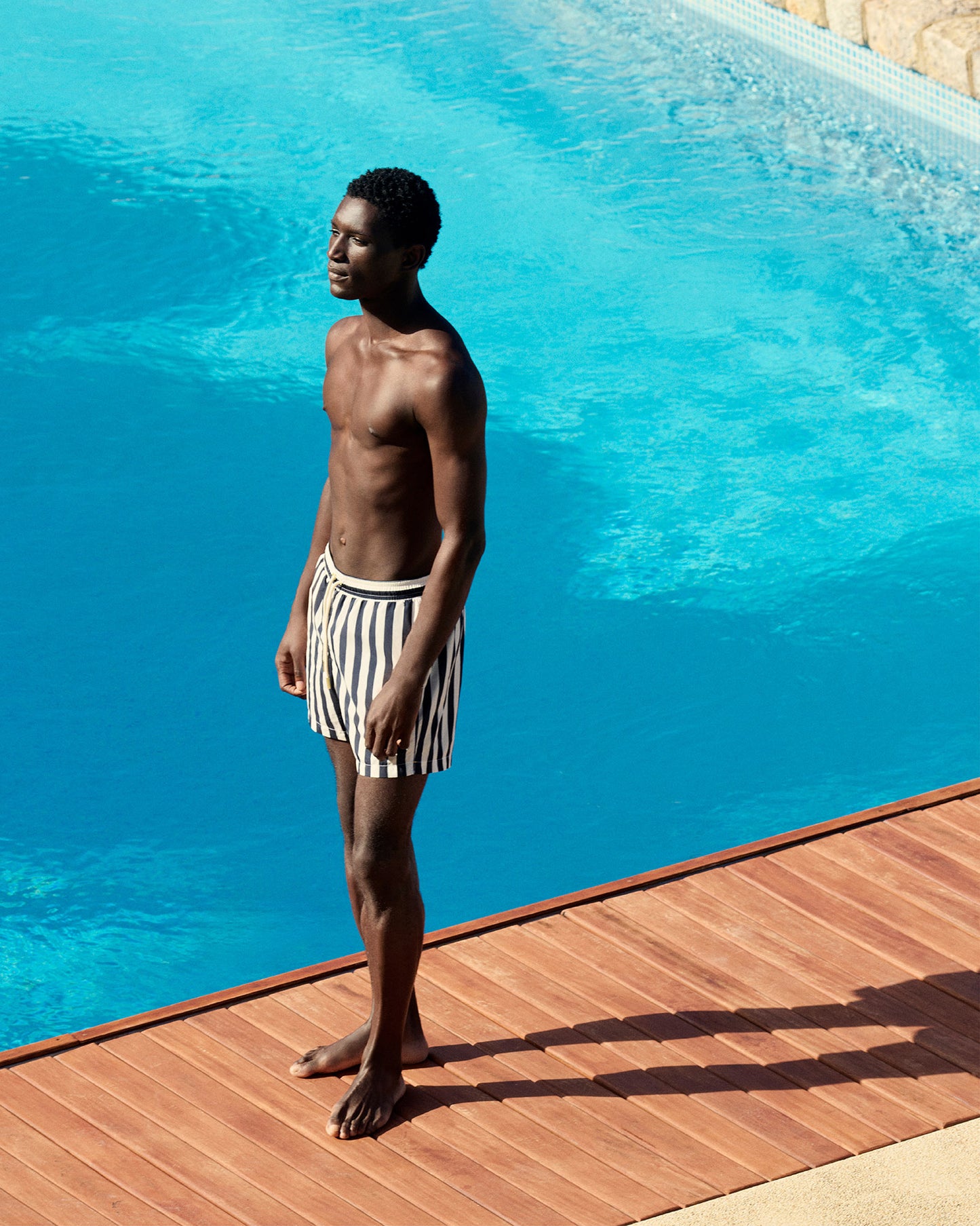 Man standing by a pool wearing striped swim shorts