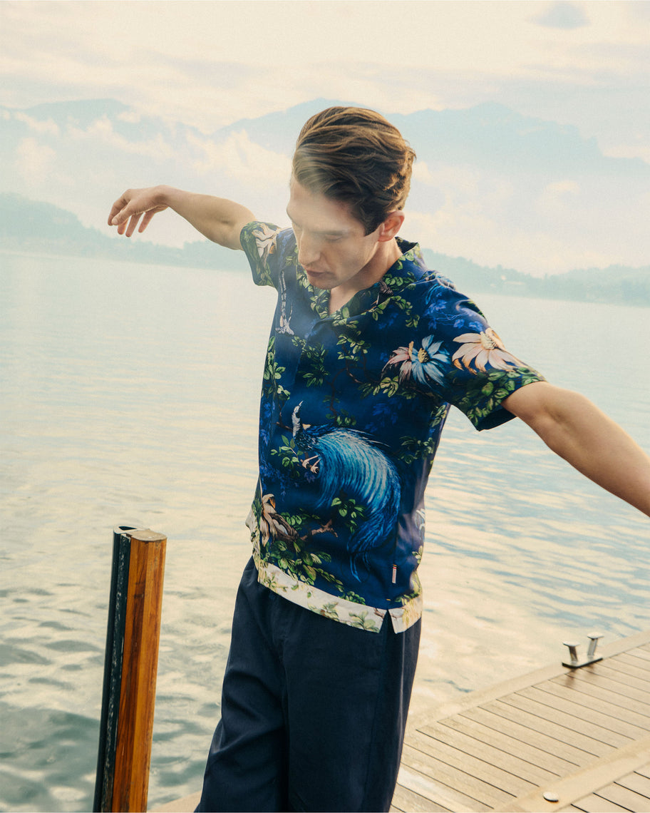 Man wearing a floral shirt by a lake with mountains in the background