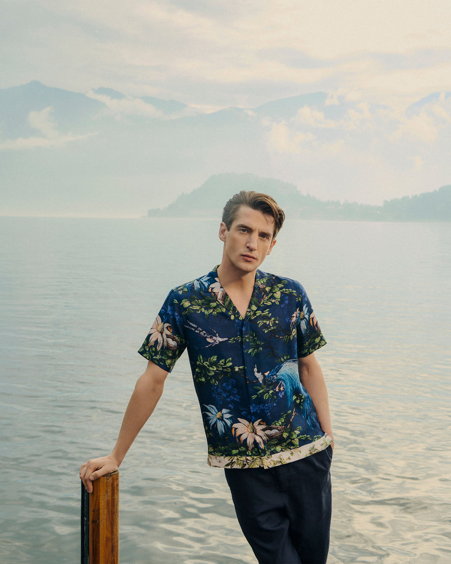 Man in a floral shirt standing by a body of water with mountains in the background