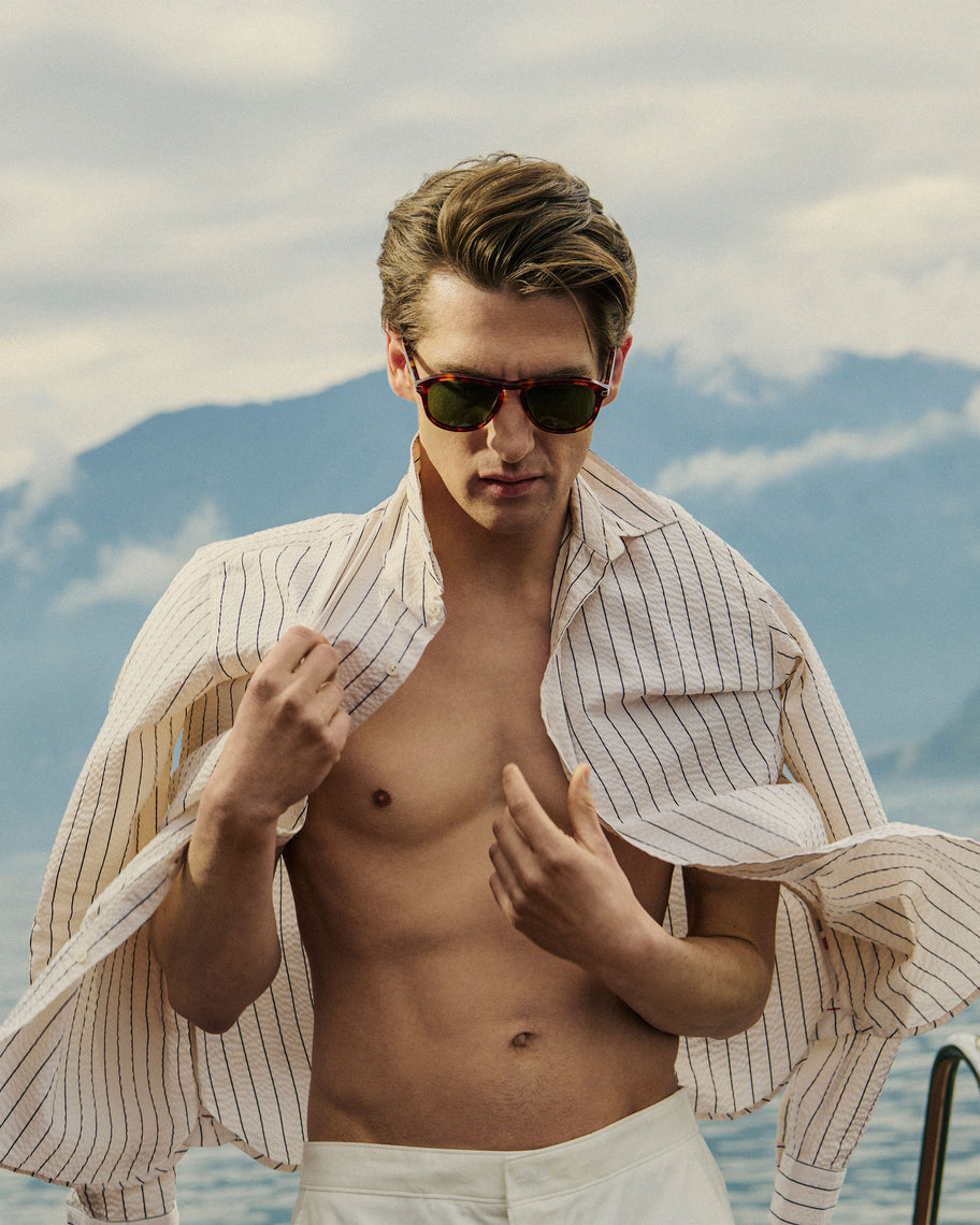 Man with sunglasses and open shirt standing by a body of water with mountains in the background